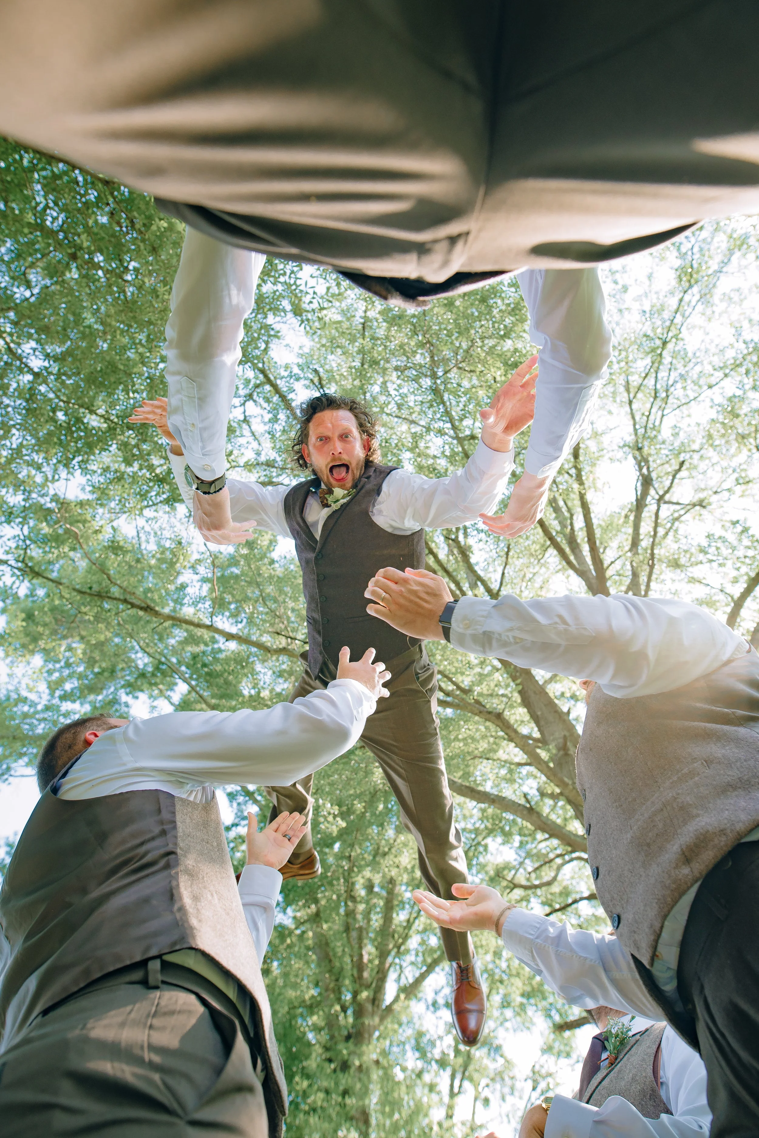A groom in a wedding suit is falling into a group of men in vests and dress shirts, with trees and sunlight overhead. Atlanta wedding. Wedding Photography by Sowing Clover Photography.