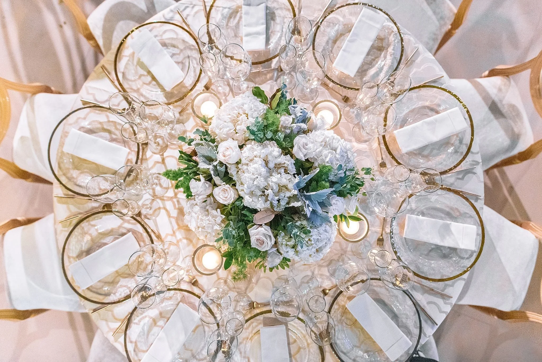 Elegant banquet table decorated with a floral centerpiece of white roses, hydrangeas, and greenery, surrounded by gold-rimmed glass chargers, white napkins, and multiple wine glasses. Nashville Wedding photography by Sowing Clover Photography.
