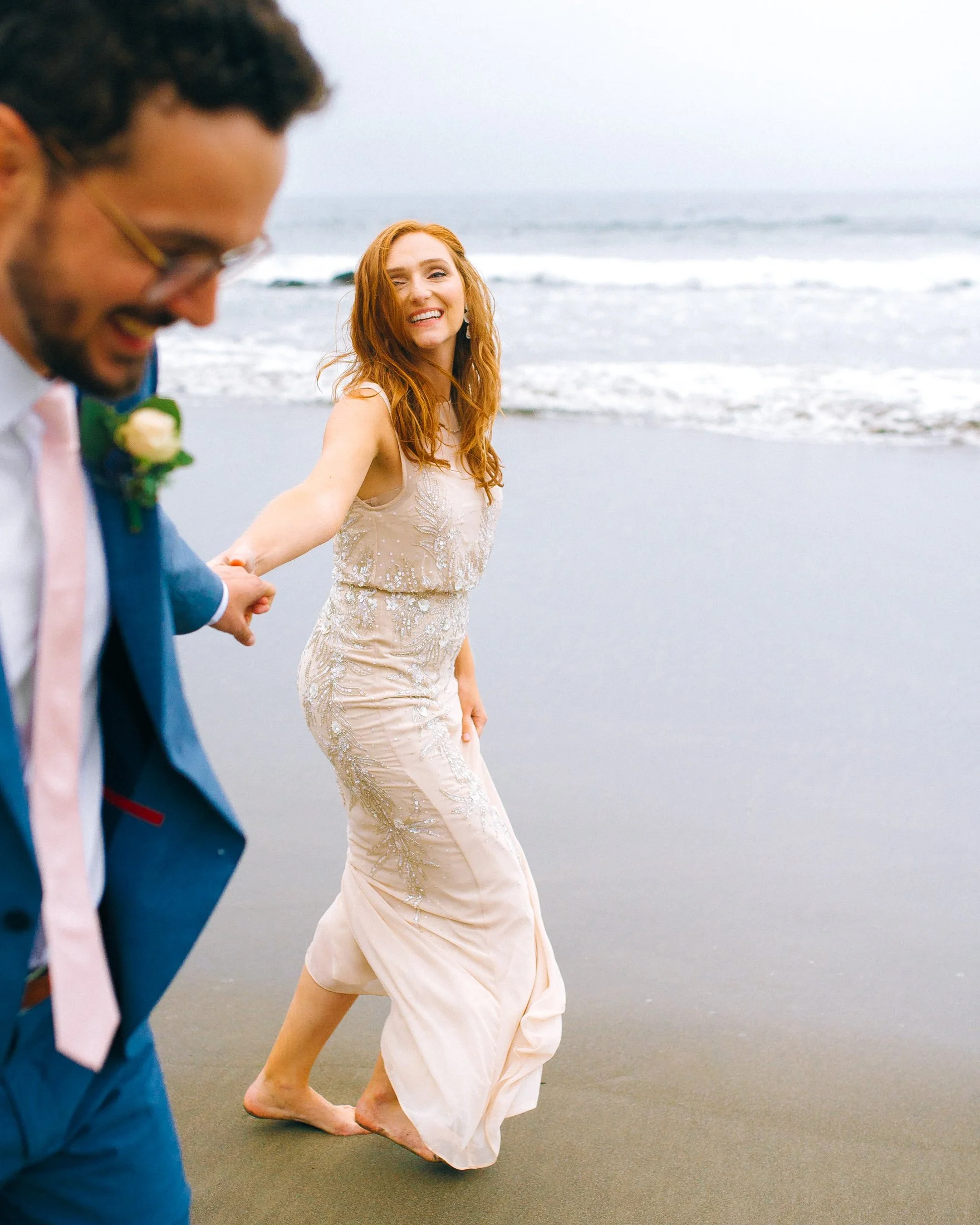 A smiling red-haired bride n in a white dress holding hands with a man in a blue suit on a beach, with ocean waves in the background in San Fransisco. Wedding Photography by Sowing Clover Photography.
