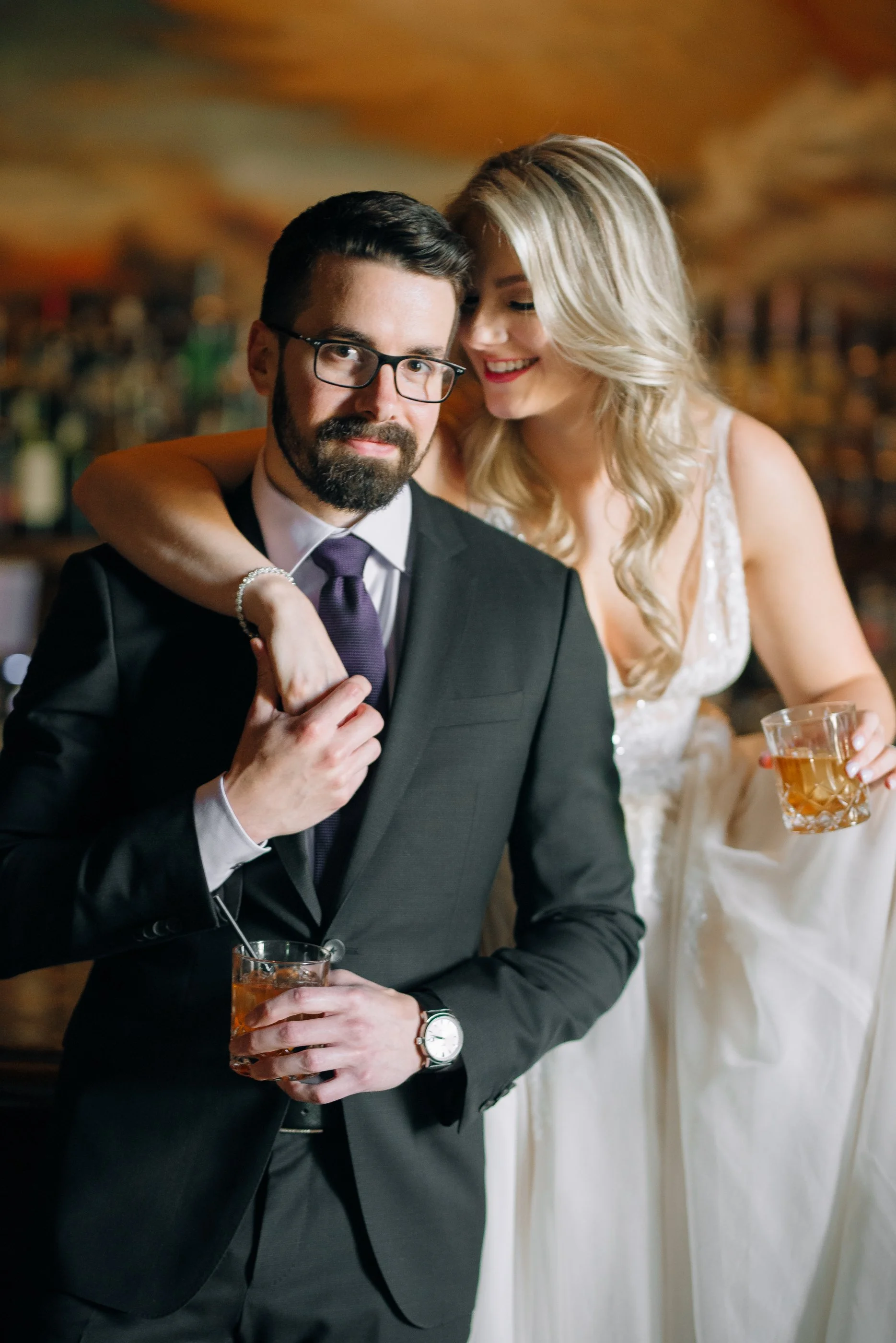 A groom with glasses with a beard, holding a glass, and a bride in a white wedding dress leaning on his shoulder with her arm around his neck, both smiling at a wedding at the Ritz in Atlanta. Wedding Photography by Sowing Clover Photography.