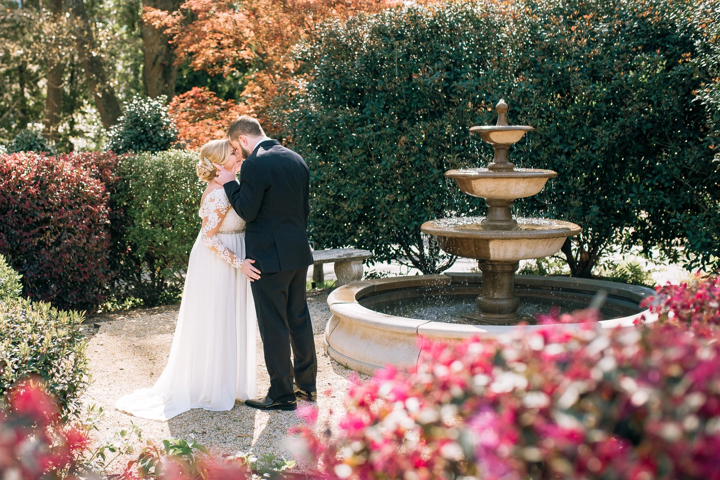 A bride and groom share a kiss next to a three-tier fountain in a garden with blooming flowers and trees at The Estate in Atlanta, GA.  Wedding Photography by Sowing Clover Photography.