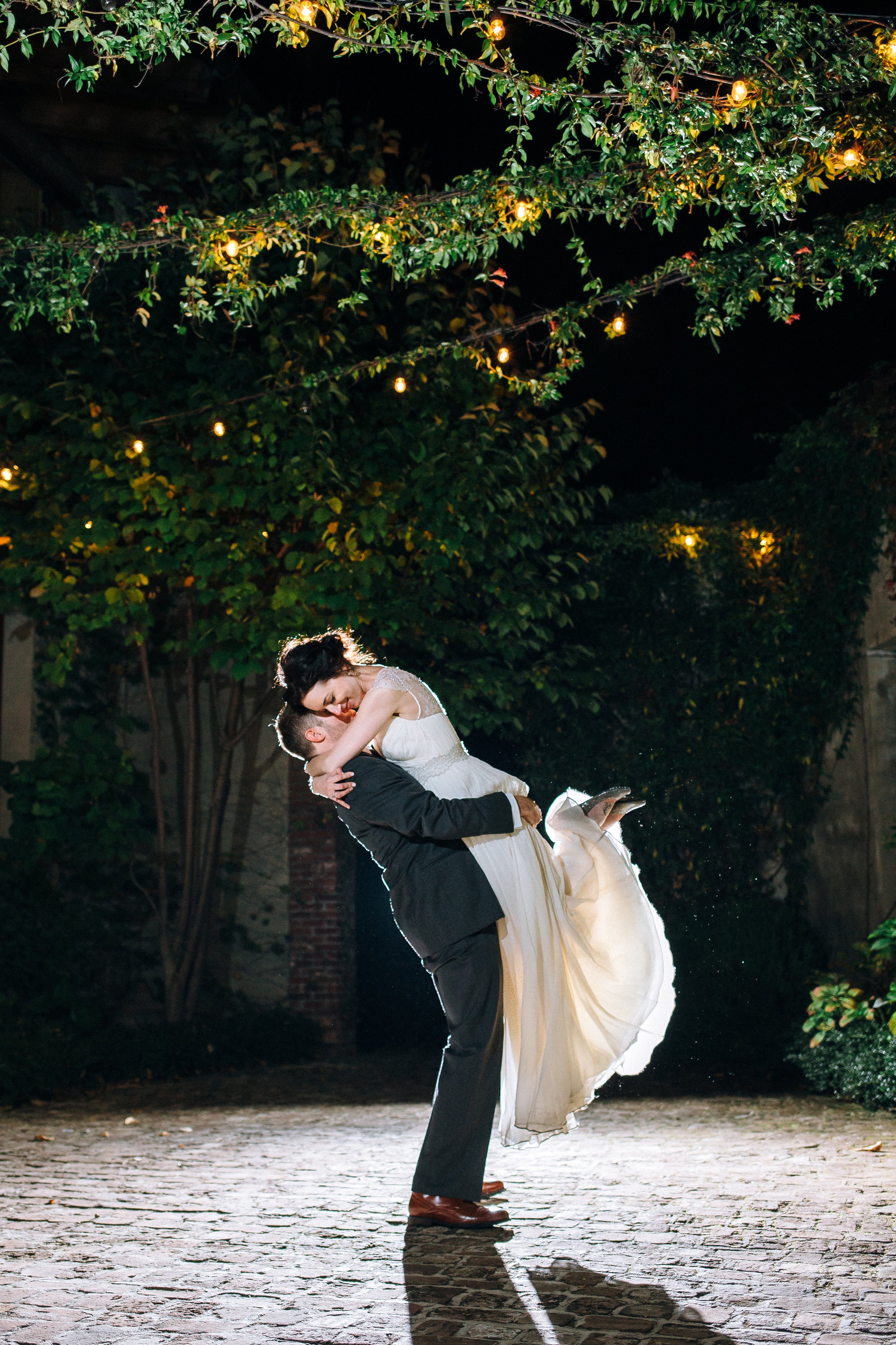 A couple dancing outdoors at night, with the man lifting the woman in a romantic pose under string lights and greenery during their wedding at summerhill studios in Atlanta