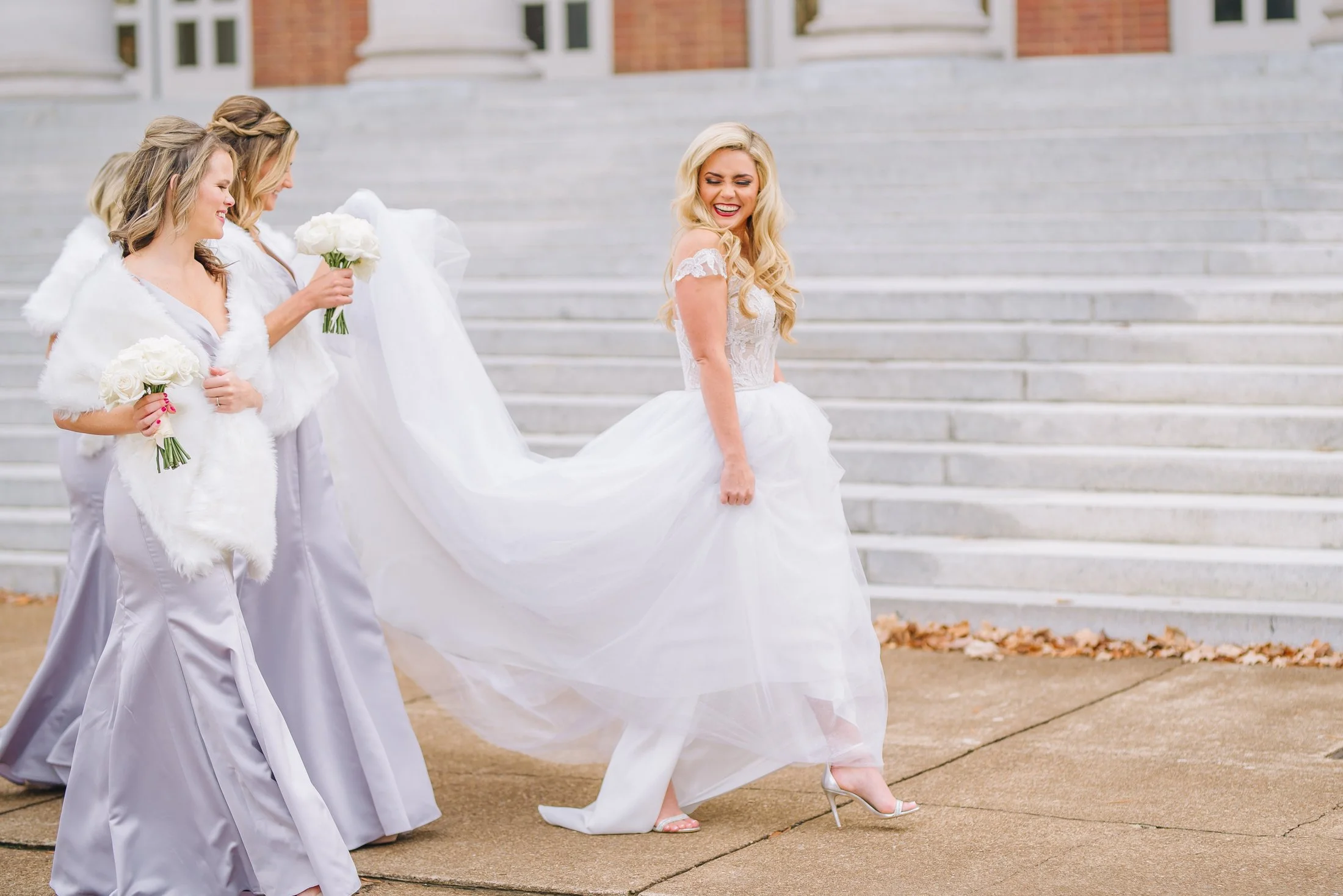 Bride in a white wedding dress with lace details, smiling and walking down stairs, accompanied by three bridesmaids holding bouquets at Vanderbilt Campus in Nashville. Wedding Photography by Sowing Clover Photography.