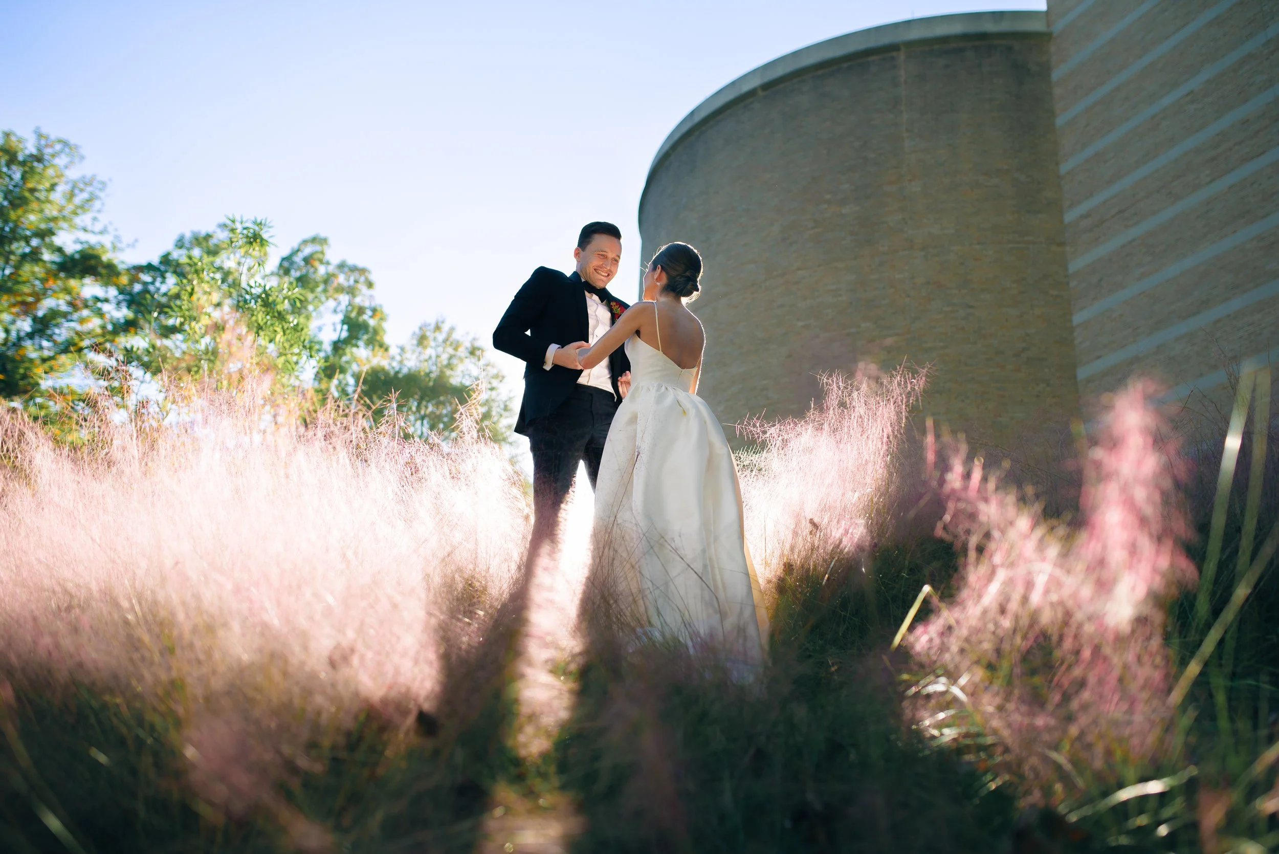 A bride and groom holding hands, smiling, outdoors during daytime with pink grass and a large brick building in the background at Fernbank Science Center. Wedding Photography by Sowing Clover Photography.
