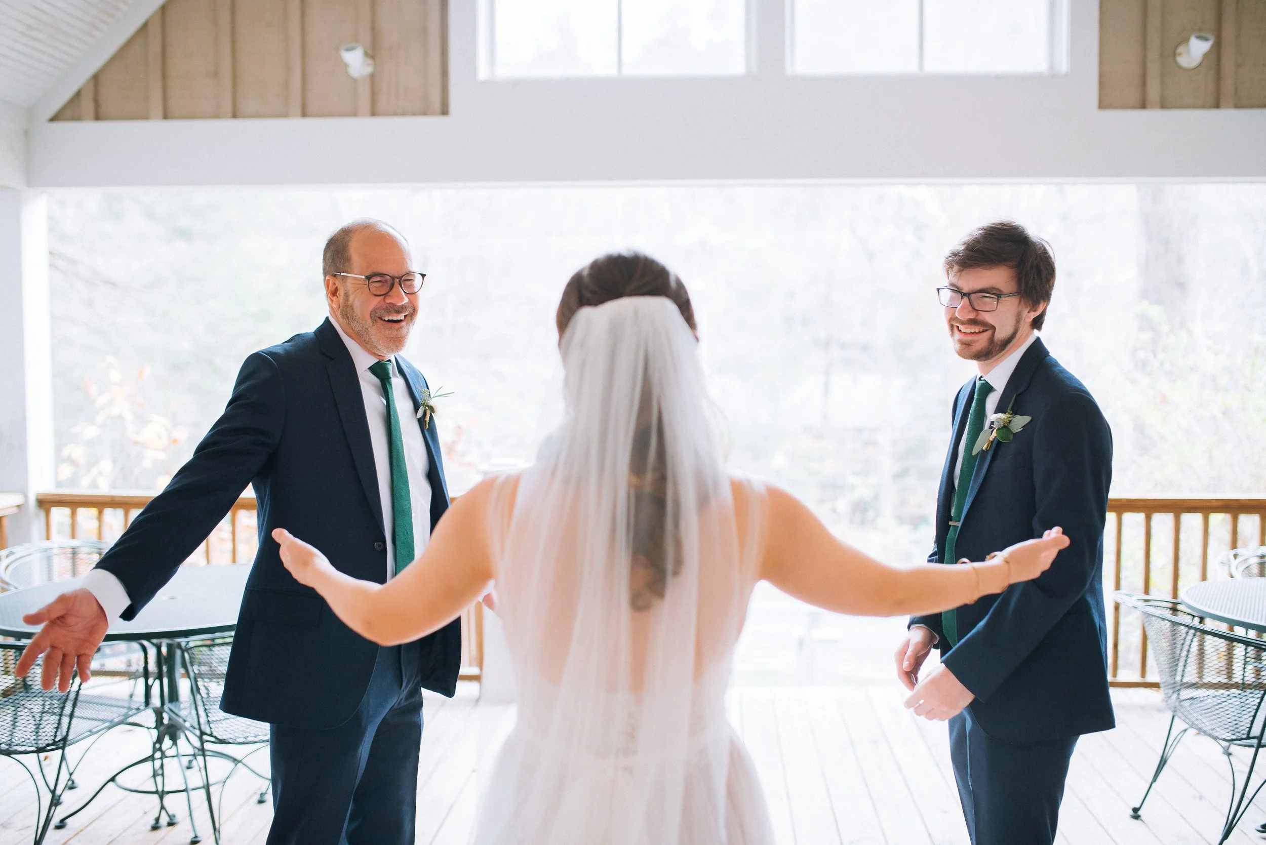 Bride with her back to the camera, arms open, first look with dad and brother in formal suits during a wedding celebration in a bright, airy hall. Montreat, NC  Wedding Photography by Sowing Clover Photography.
