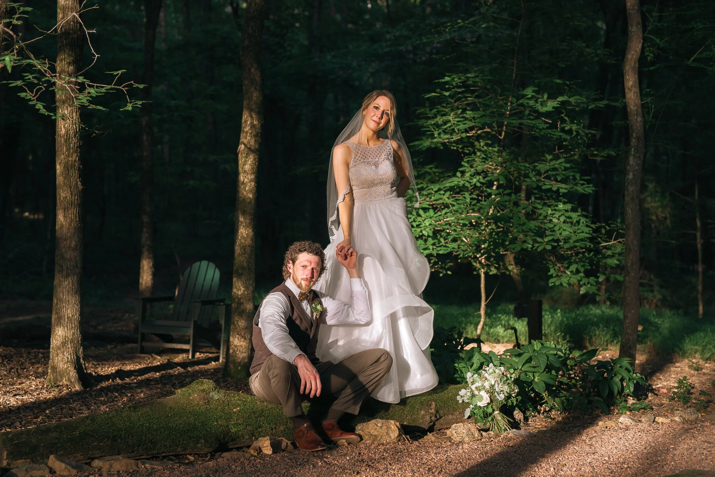 A bride and groom in wedding attire posing outdoors in a wooded area during daytime. The bride is standing on a moss-covered rock, holding hands with the groom, who is sitting on the ground. Atlanta wedding. Wedding Photography by Sowing Clover Photo