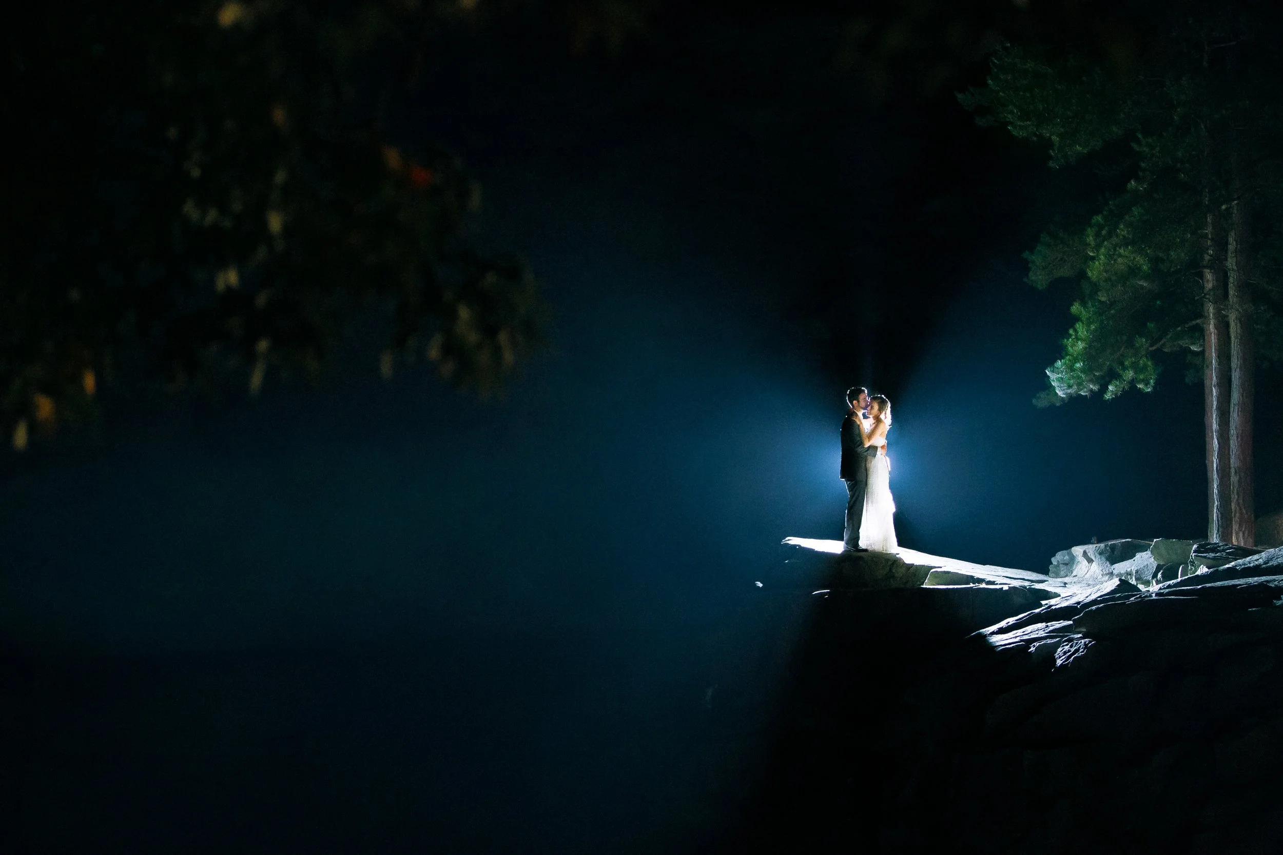 A couple in wedding attire standing on a rocky ledge at night, illuminated by a bright light with dark trees in the background in Nashville TN Wedding photography by Sowing Clover Photography