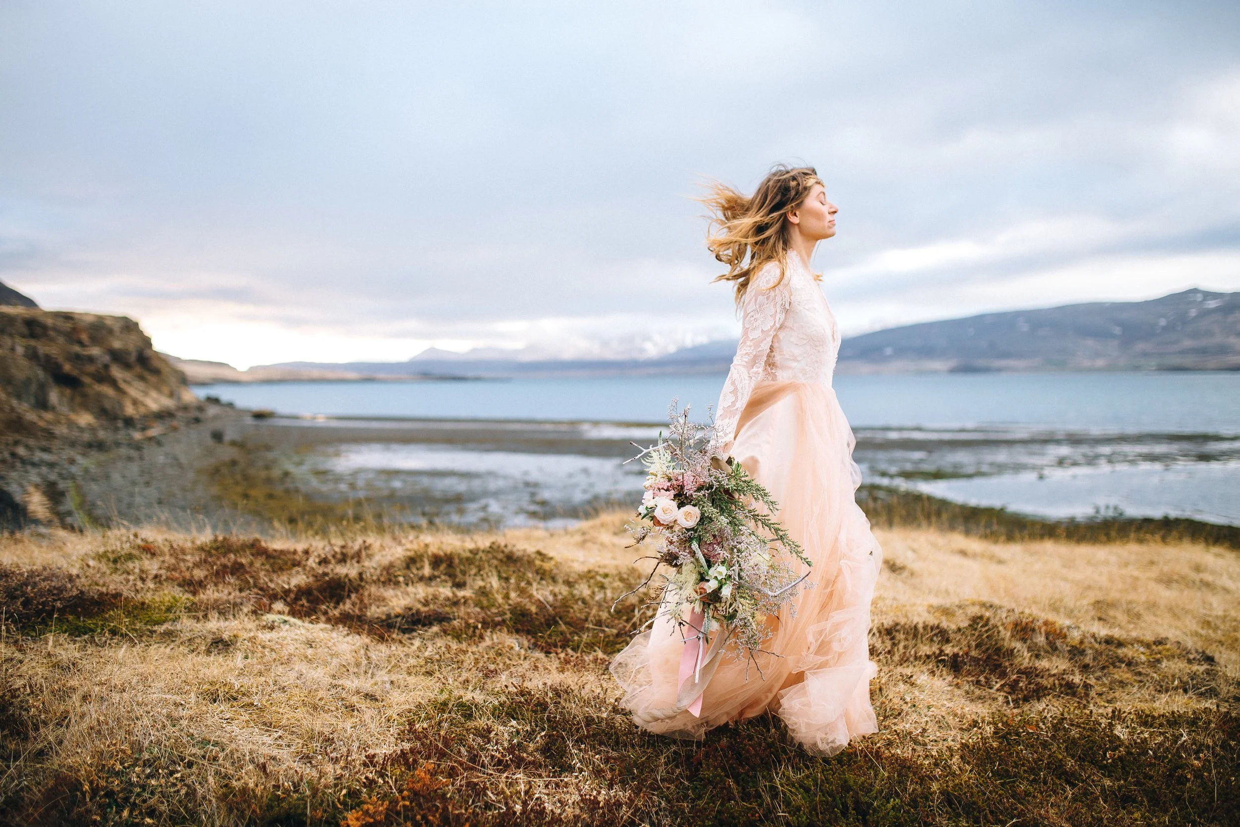 A woman in a lace wedding dress holding a large bouquet of flowers stands on a rocky, grassy terrain near a body of water with mountains in the background in Iceland. Wedding Photography by Sowing Clover Photography.