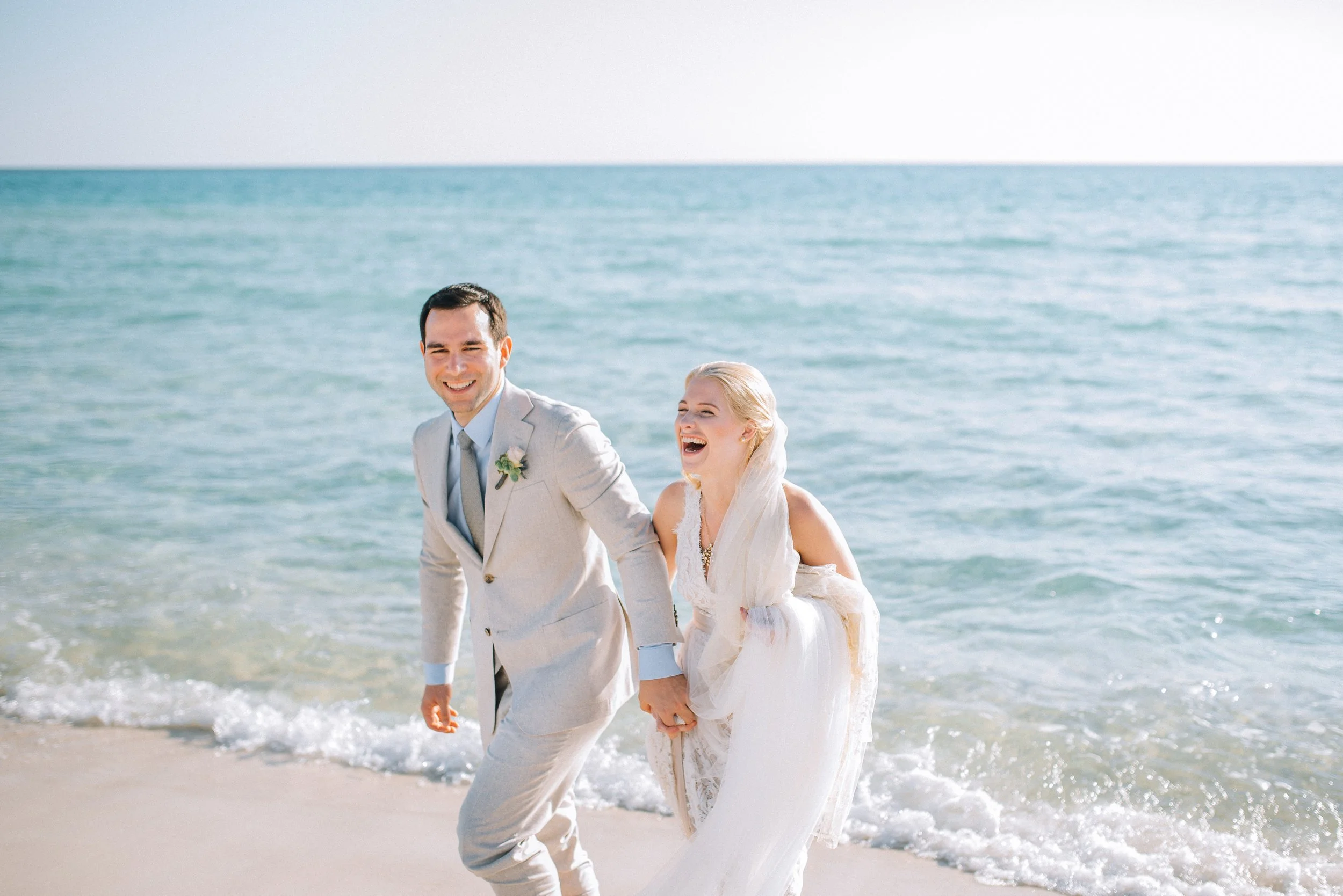 A newlywed couple in wedding attire holding hands and laughing on a beach in Florida. Wedding Photography by Sowing Clover Photography.