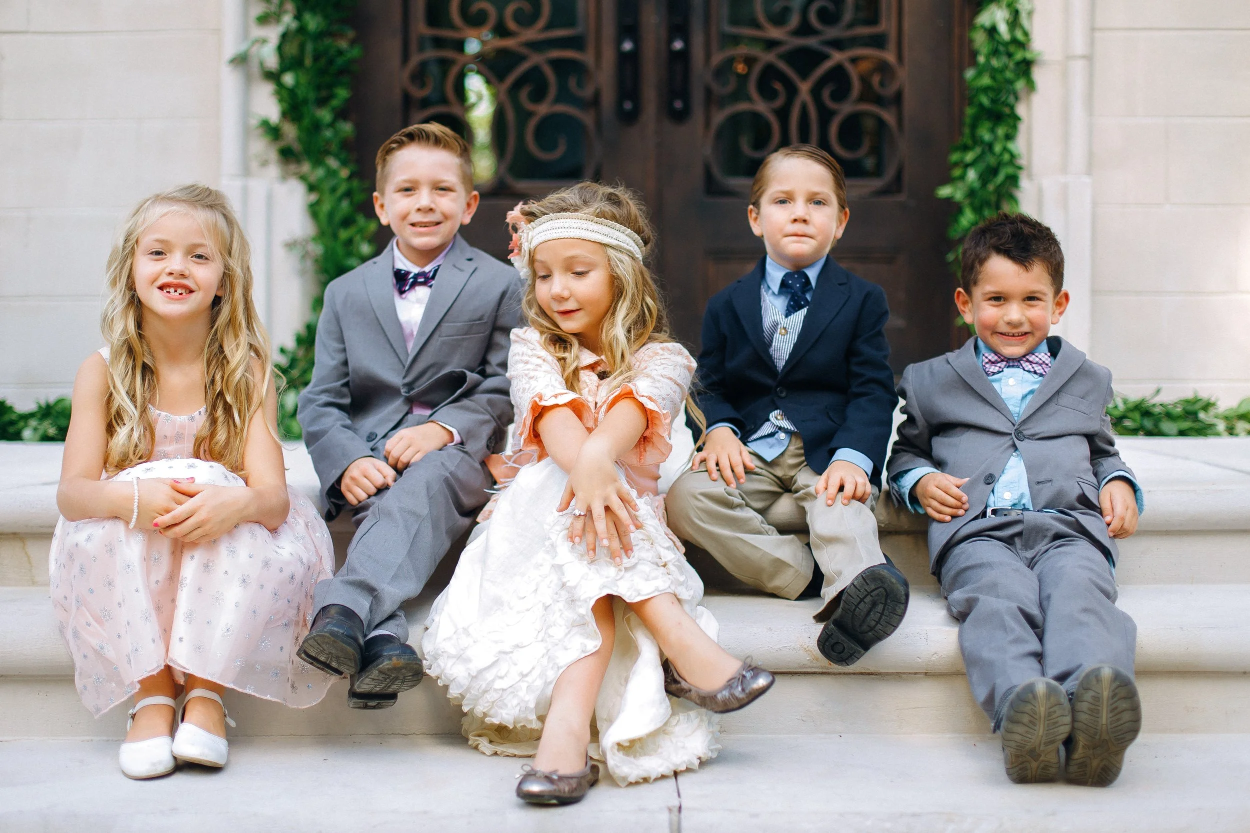 Five young children sitting on steps in front of a large wooden door, dressed in formal clothing for a wedding ceremony in Atlanta Georgia. Wedding photography by Sowing Clover Photography.