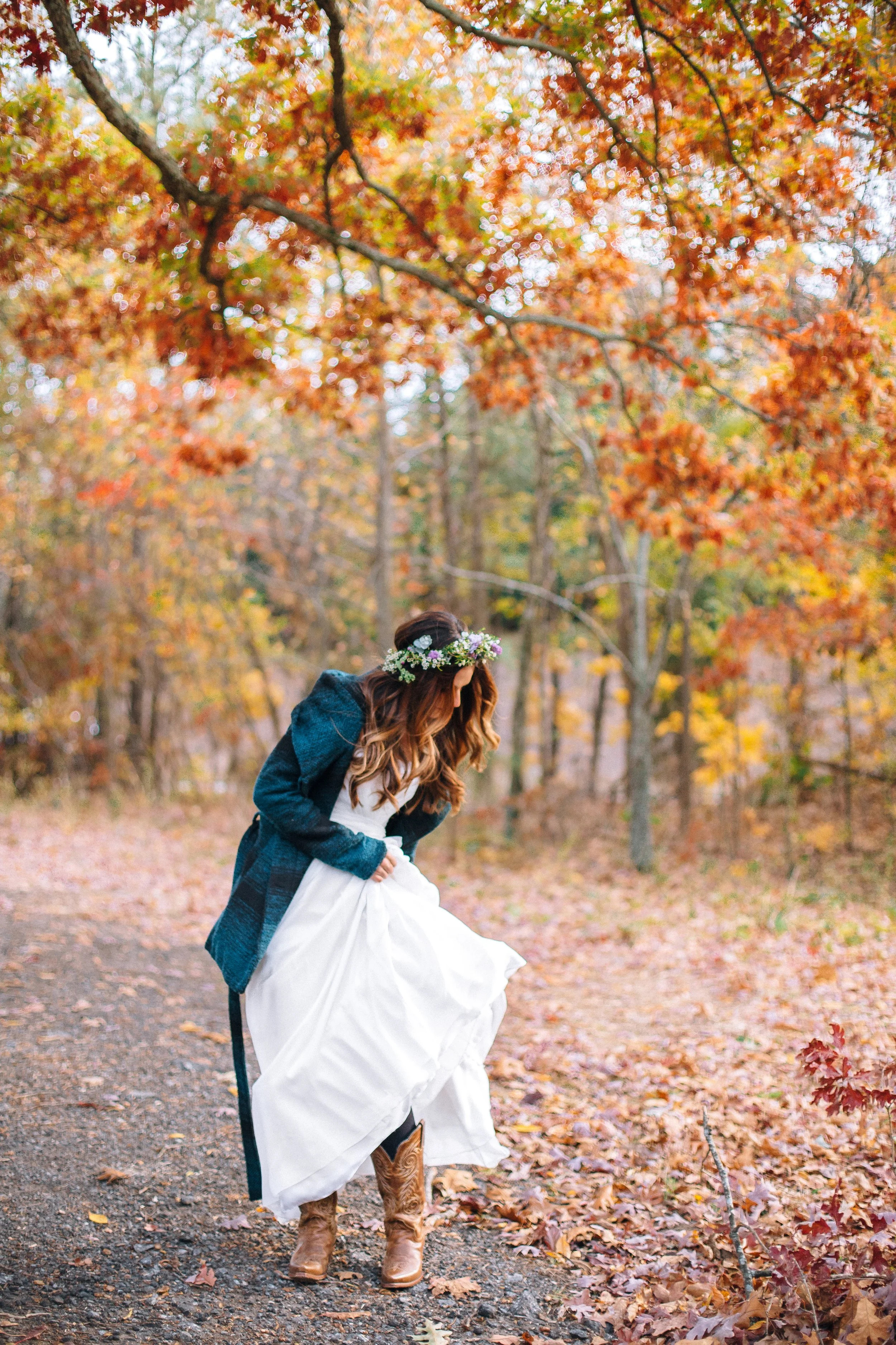 A bride in fall clothing, a white wedding dress, brown boots, and a floral crown, standing on a wooded path with autumn leaves and trees with orange and yellow foliage in Atlanta.  Wedding Photography by Sowing Clover Photography.