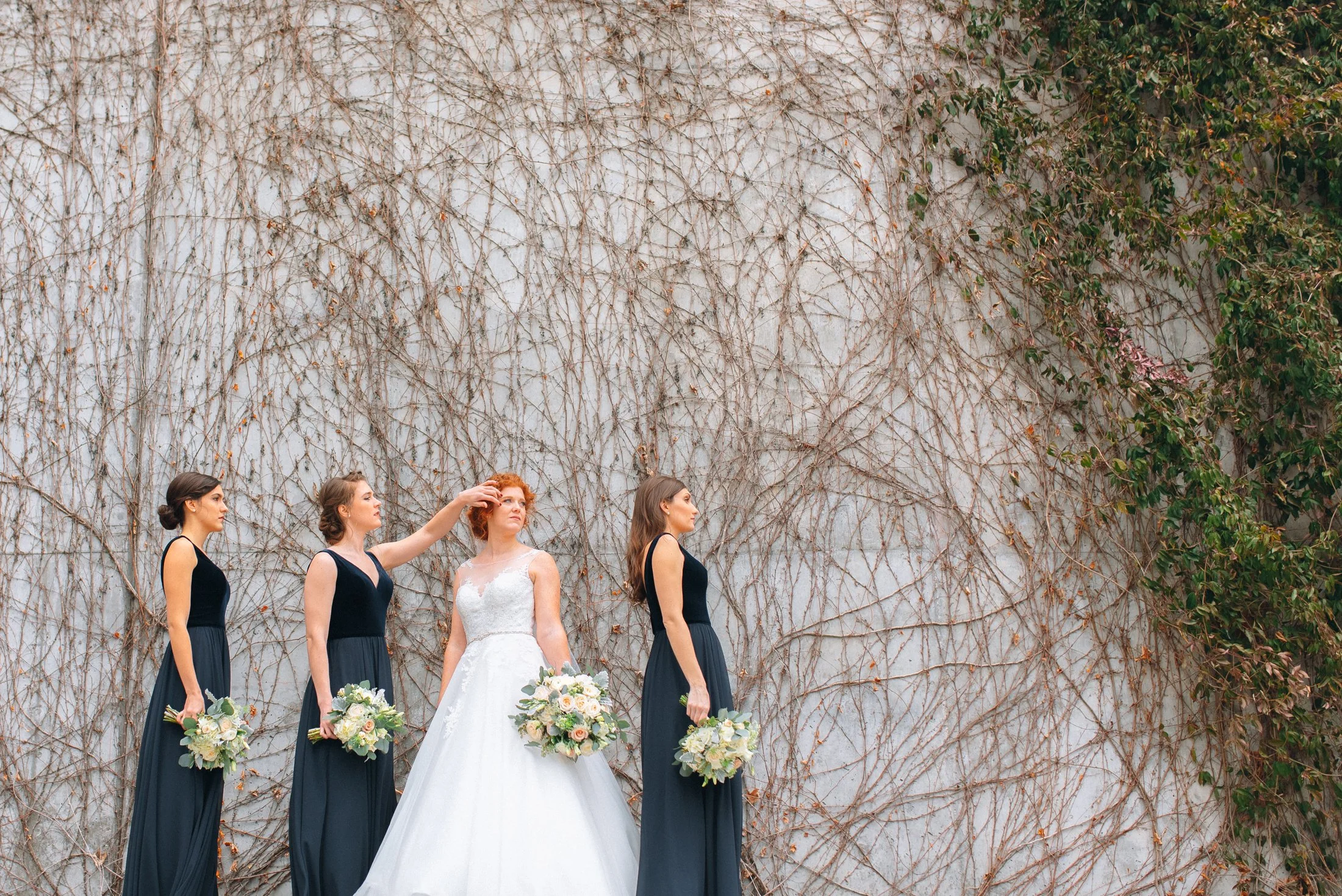 Wedding bride in white dress with three bridesmaids in navy dresses holding bouquets against a vine-covered wall at Hermitage hotel in Nashville TN. Wedding Photography by Sowing Clover Photography.