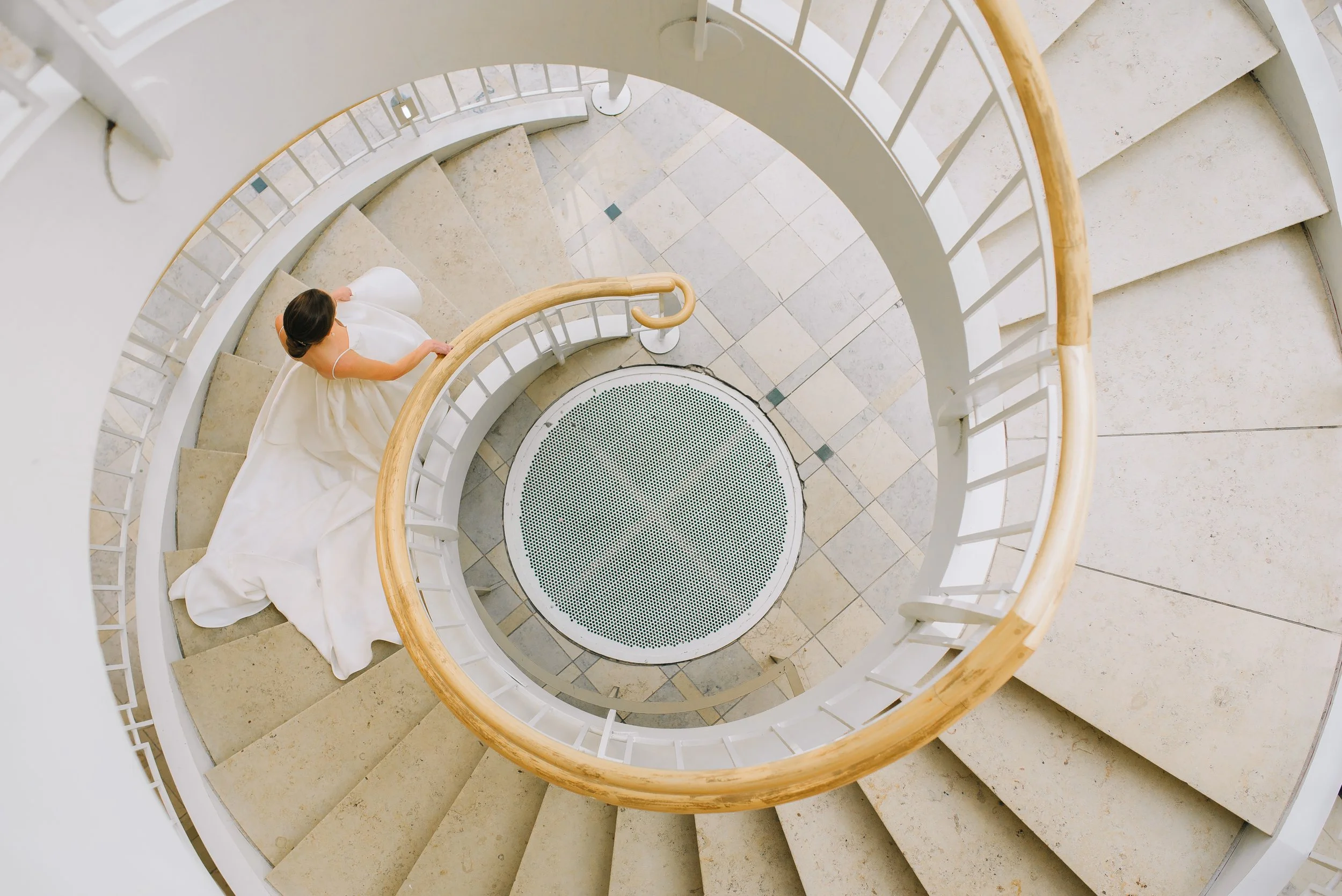 A bride in a white dress walking down a spiral staircase, seen from above at the Fernbank Science Center. Wedding Photography by Sowing Clover Photography.