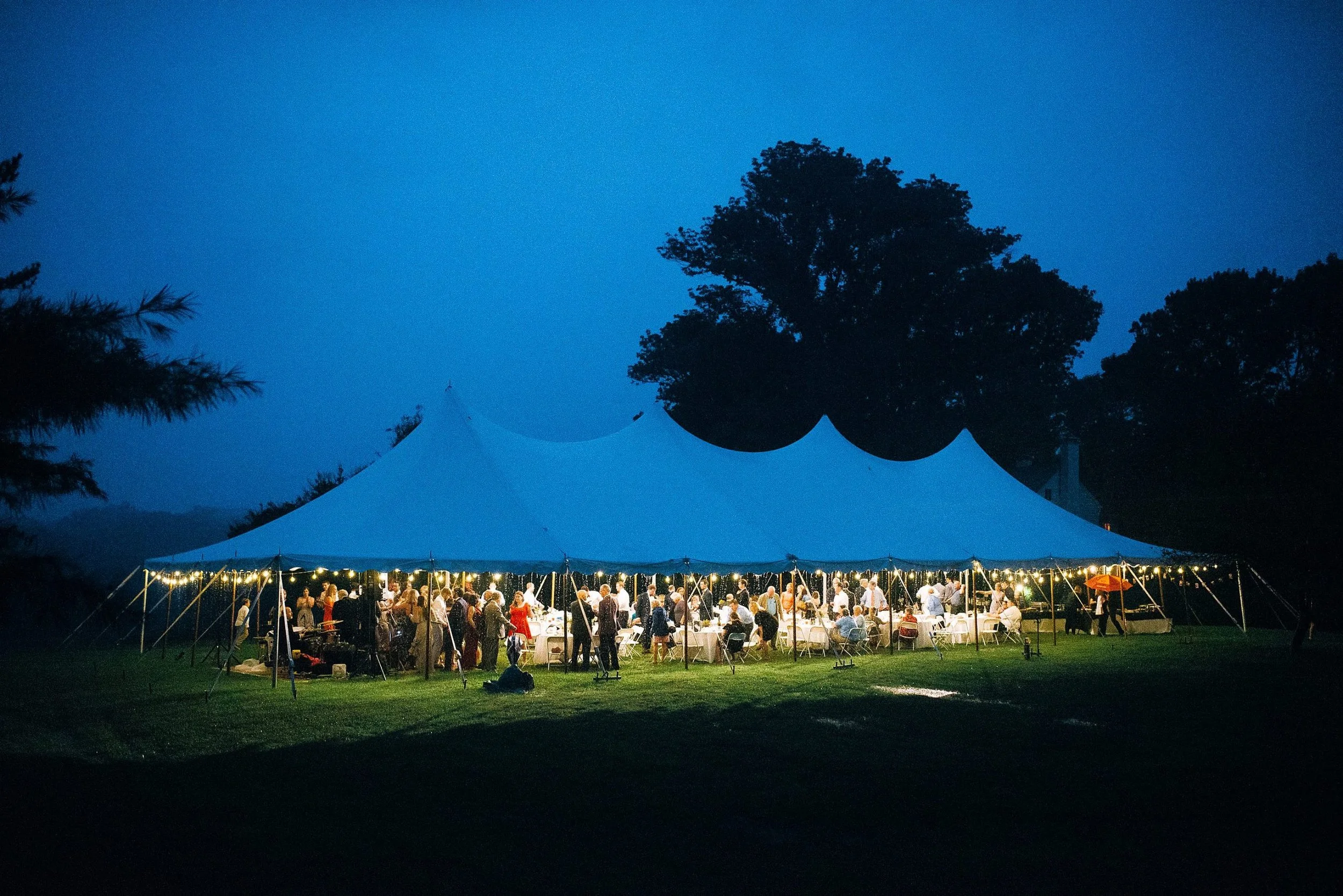 A large outdoor wedding reception under a white tent at dusk with string lights, surrounded by trees and grassy area, with people gathered inside and outside in Nashville TN.  Wedding photography by Sowing Clover Photography