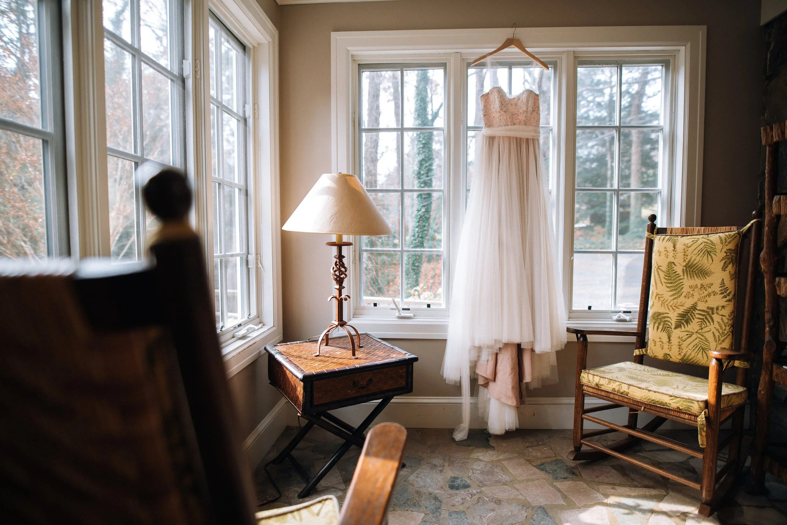 A white wedding dress hanging in front of a window in a cozy room with vintage furniture and a stone floor. Montreat North Carolina. photography by Sowing Clover Photography.