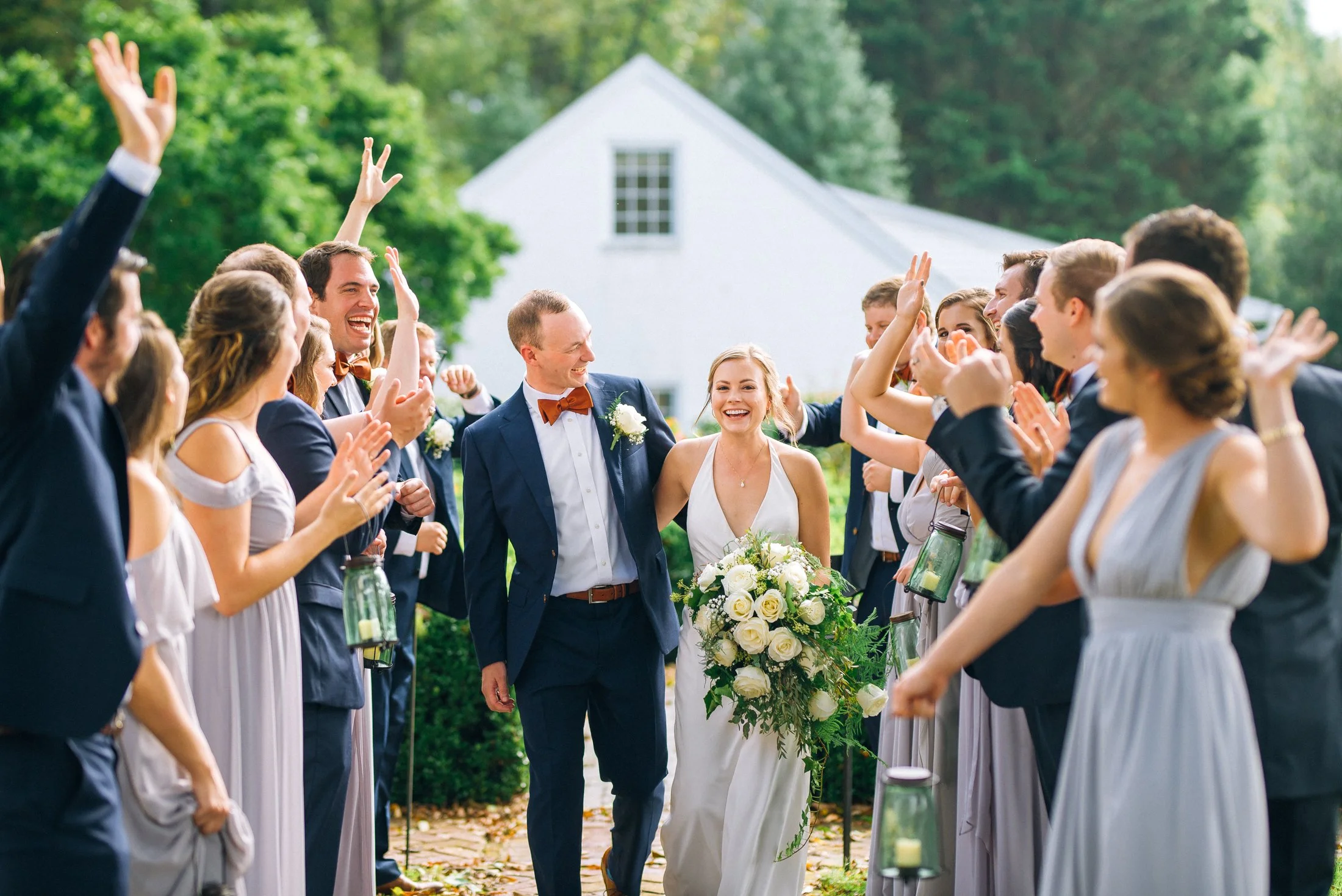 Bride and groom walking through a crowd of wedding guests outside, surrounded by trees and greenery, celebrating the wedding. Wedding Photography by Sowing Clover Photography.