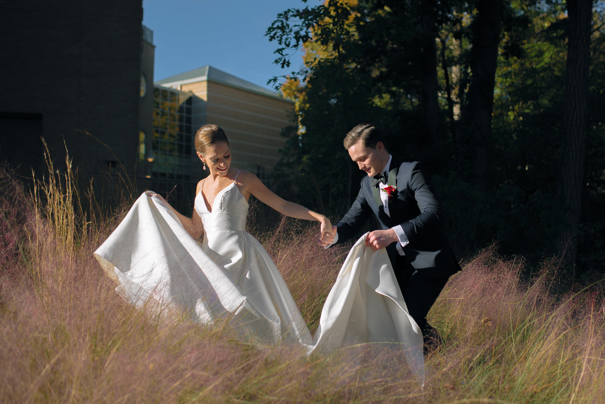 Bride and groom dancing outdoors in a field of tall grass during daytime, holding hands and smiling, with trees and modern buildings in the background at Fernbank Science Center. Wedding Photography by Sowing Clover Photography.