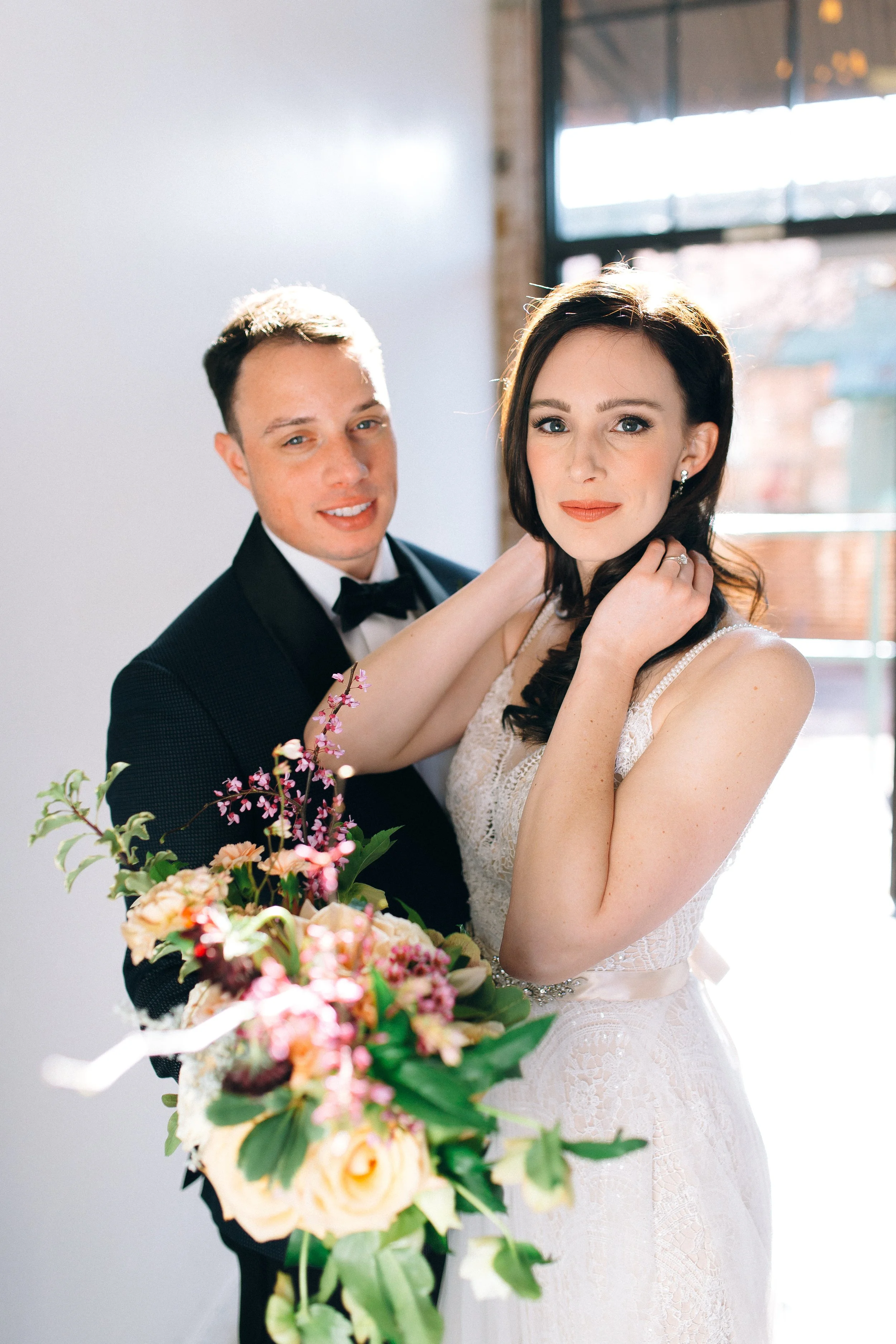 A bride in a lace wedding dress holding a bouquet of flowers and a groom in a tuxedo standing beside her indoors with sunlight streaming in. Magazine Style Photo at King Plow in Atlanta, Georgia. Wedding Photography by Sowing Clover Photography.