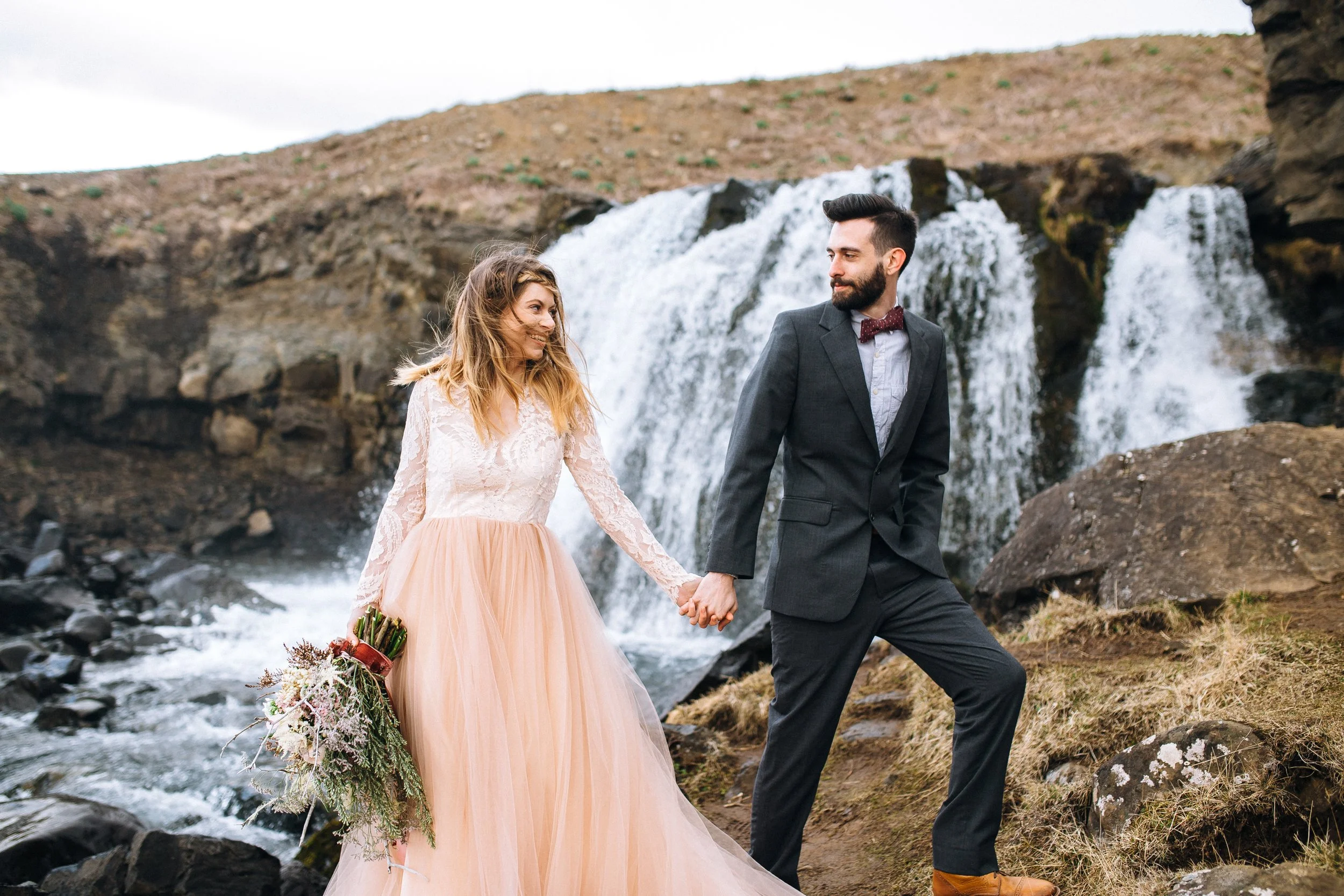 A bride and groom holding hands near a waterfall, with the bride wearing a lace dress and holding a floral bouquet, and the groom in a dark suit and bow tie, outdoors  in Iceland. Wedding Photography by Sowing Clover Photography..