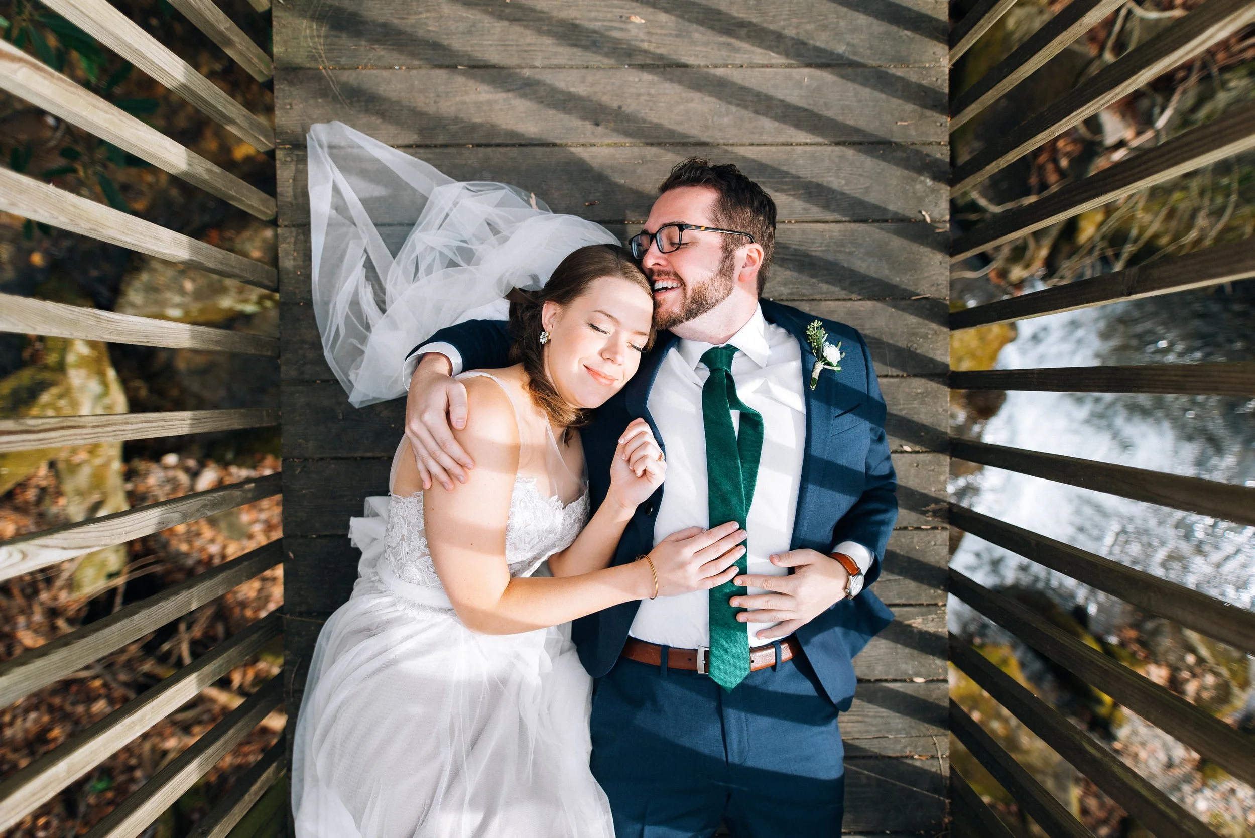 A bride and groom lying on a wooden bridge, smiling and embracing each other, enjoying a moment together on their wedding day for their wedding photography in Montreal North Carolina