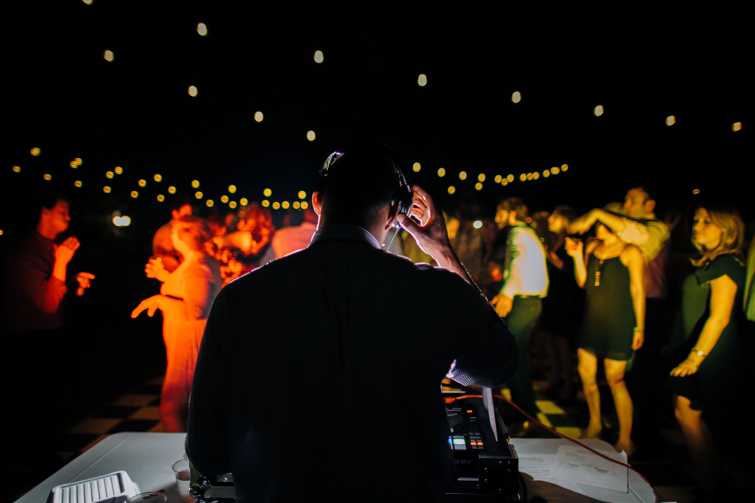 Dj wearing headphones and adjusting them while people dance at wedding reception under string lights at night. Atlanta Georgia Wedding. Wedding Photography in by Sowing Clover Photography