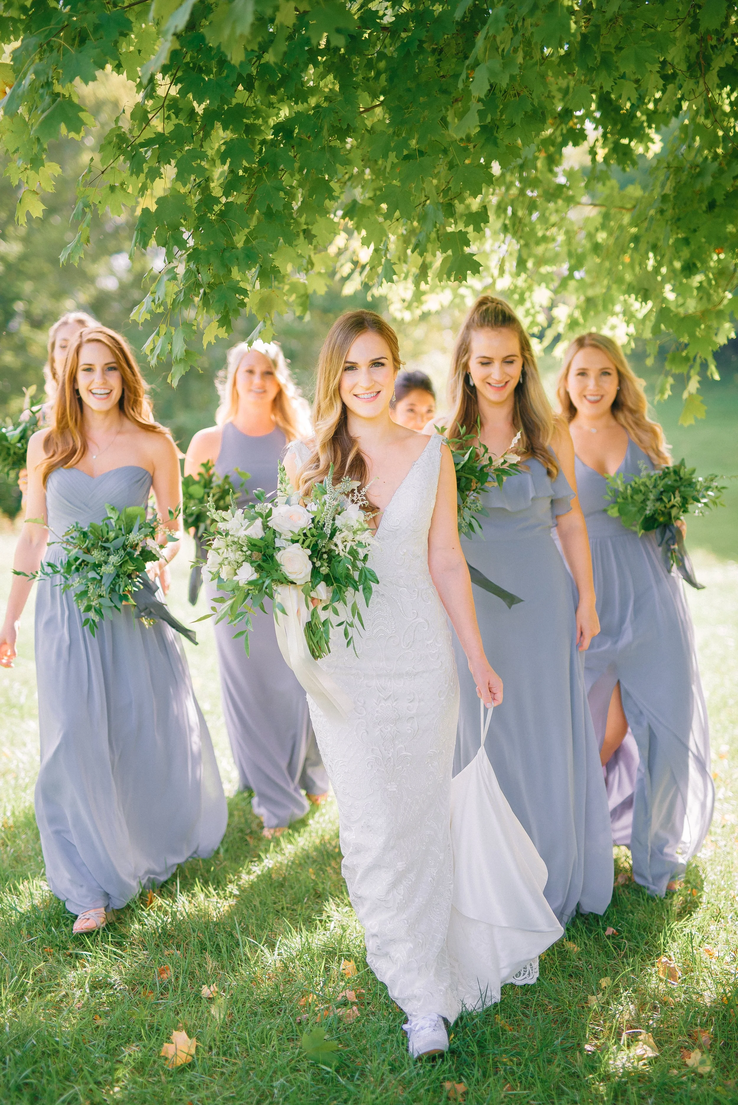 A bride walking outdoors with her bridesmaids under a leafy tree, all holding bouquets and smiling happily at Mint Springs Farm in Nashville, TN. Wedding Photography by Sowing Clover Photography.