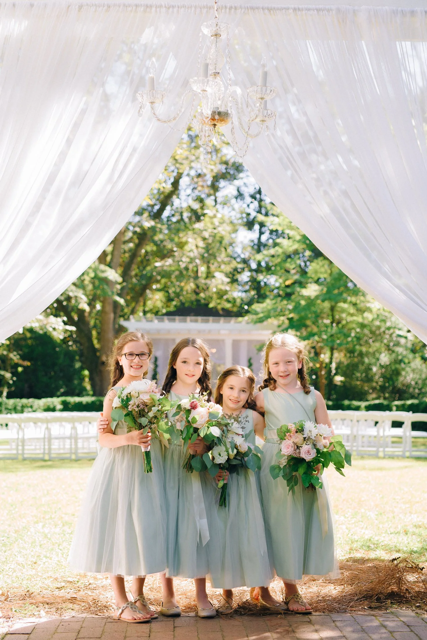 Four young flower girls in matching light green dresses holding bouquets of flowers standing under a white canopy in Savannah GA for a wedding.  Wedding Photography by Sowing Clover Photography.