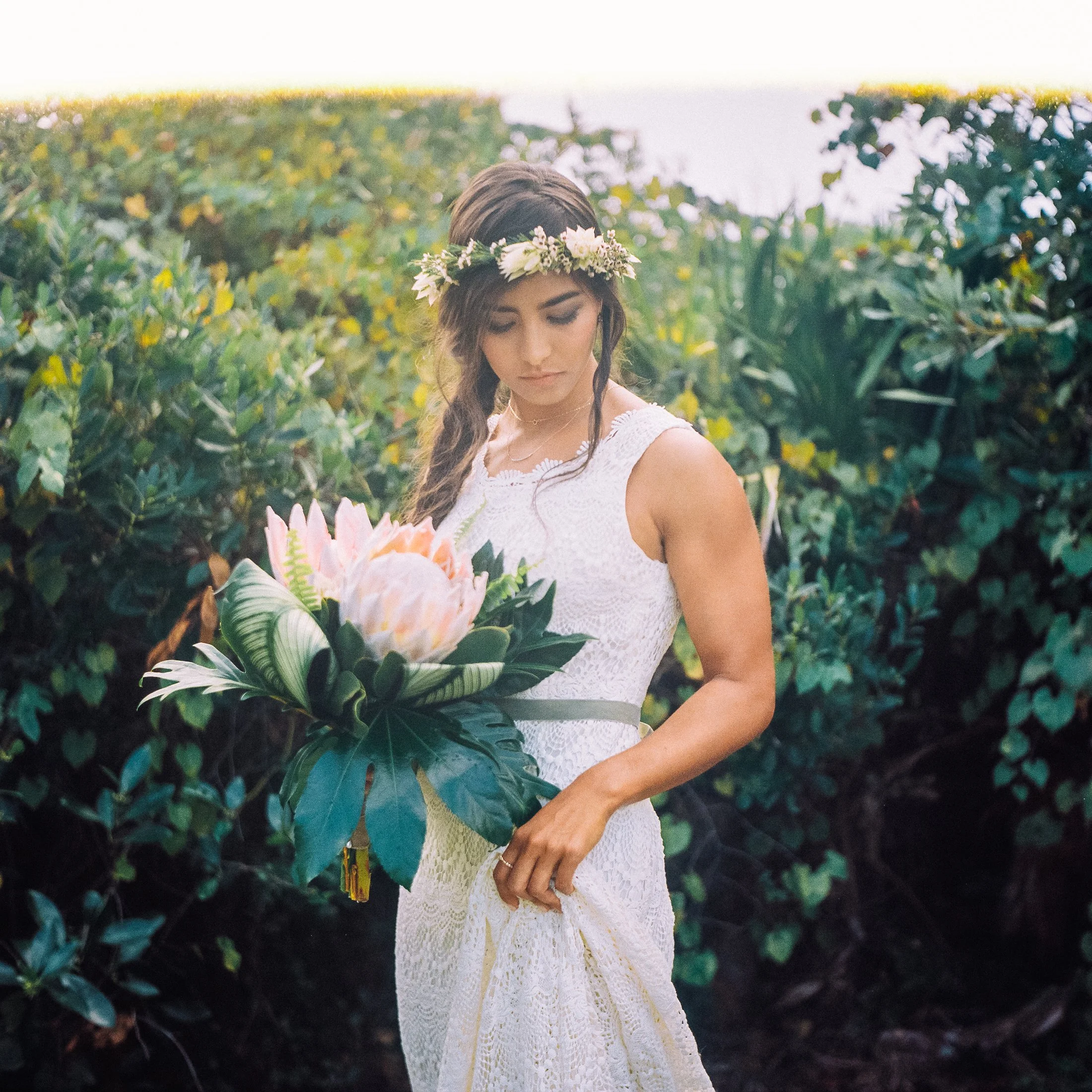 Young bride woman with long wavy hair wearing a white lace dress and floral crown, holding a bouquet of pink flowers and greenery, standing outdoors among green foliage in a wedding dress Wedding Photography in Florida by Sowing Clover Photography
