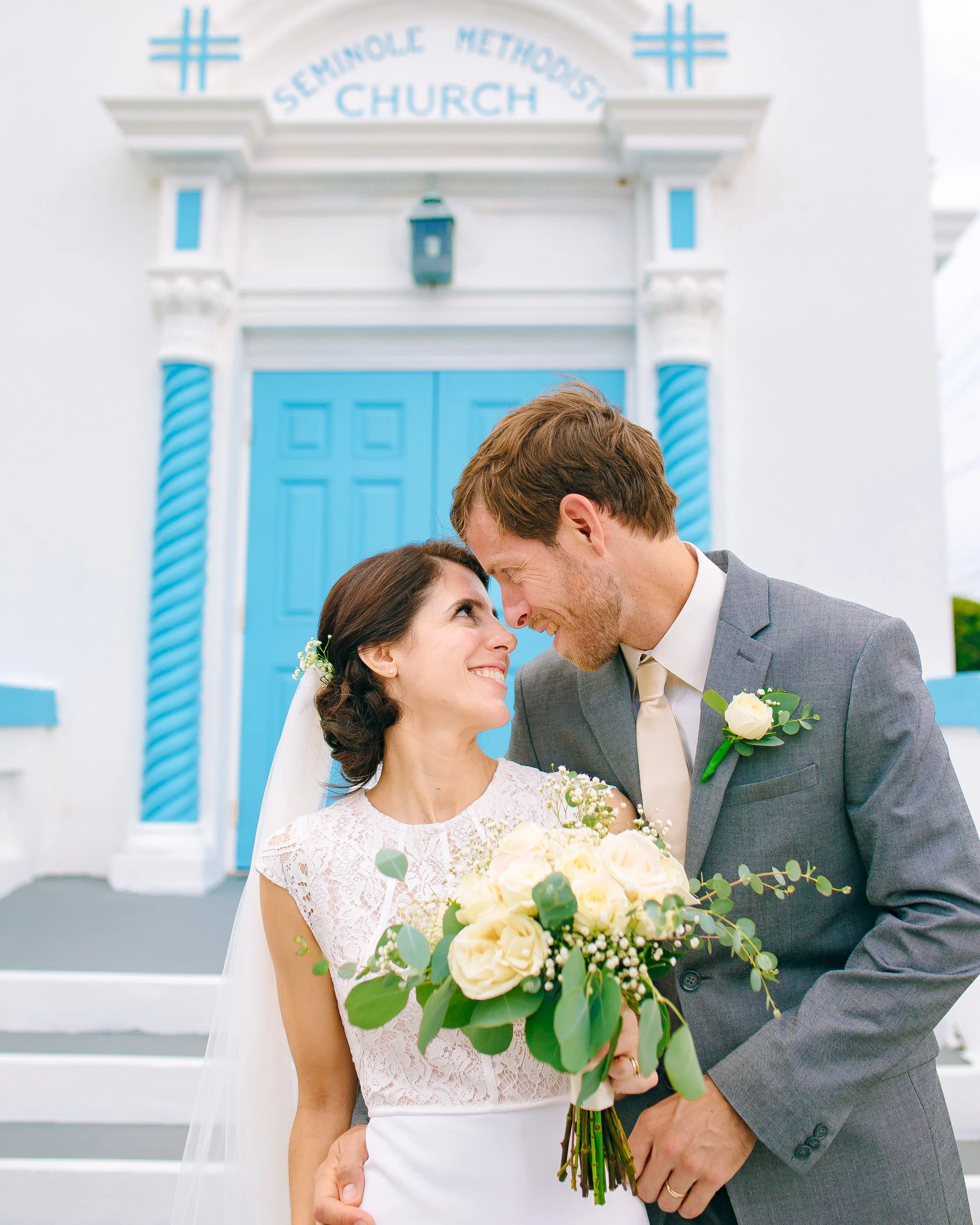 A bride and groom smiling with heir foreheads together in front of a blue door at a church, with the bride holding a bouquet of white roses, outside a white Presbyterian church. Wedding Photography by Sowing Clover Photography.