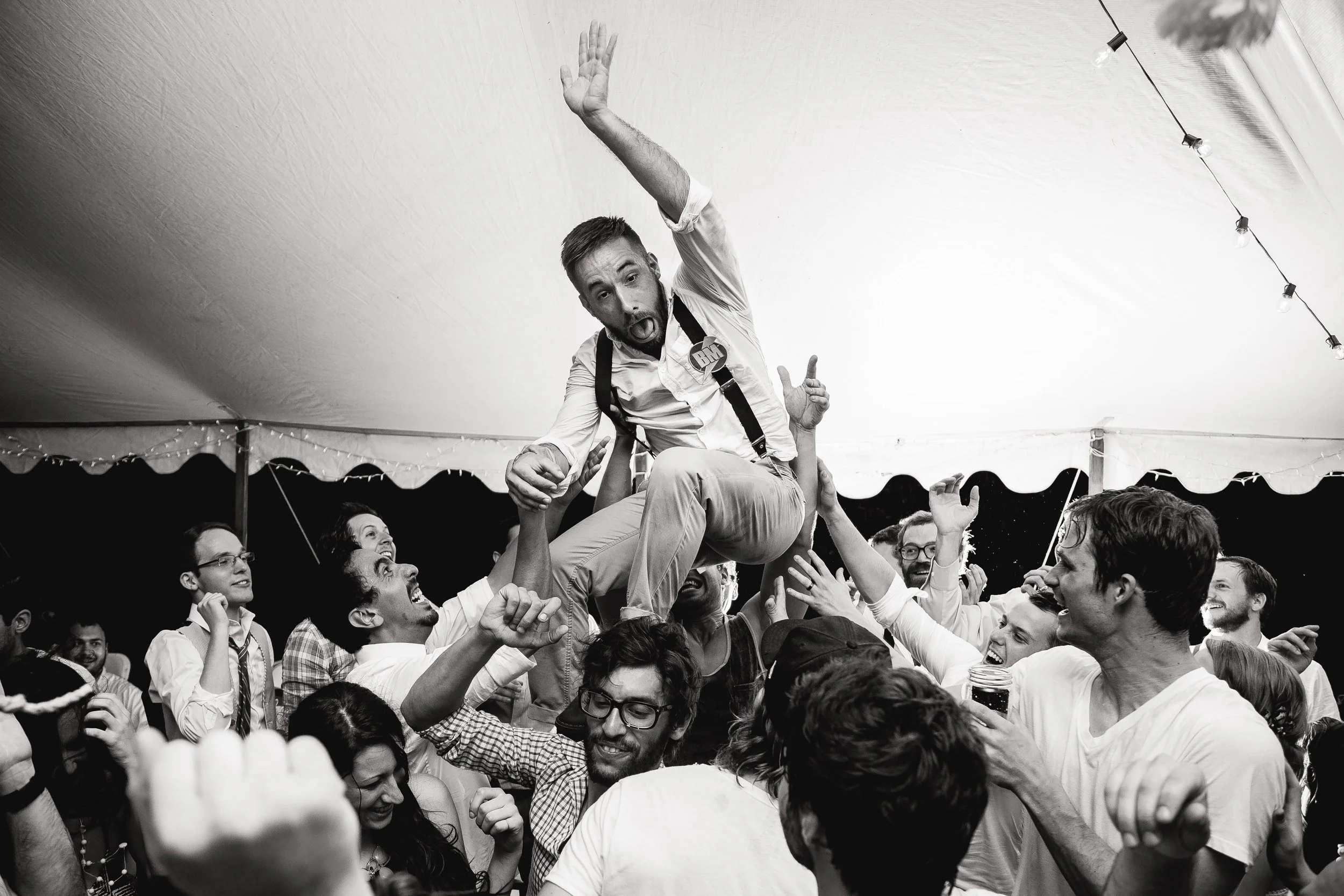 A man is crowd surfing at a lively wedding reception surrounded by smiling and cheering people under a large tent with string lights. North Georgia Wedding photography by Sowing Clover Photography.