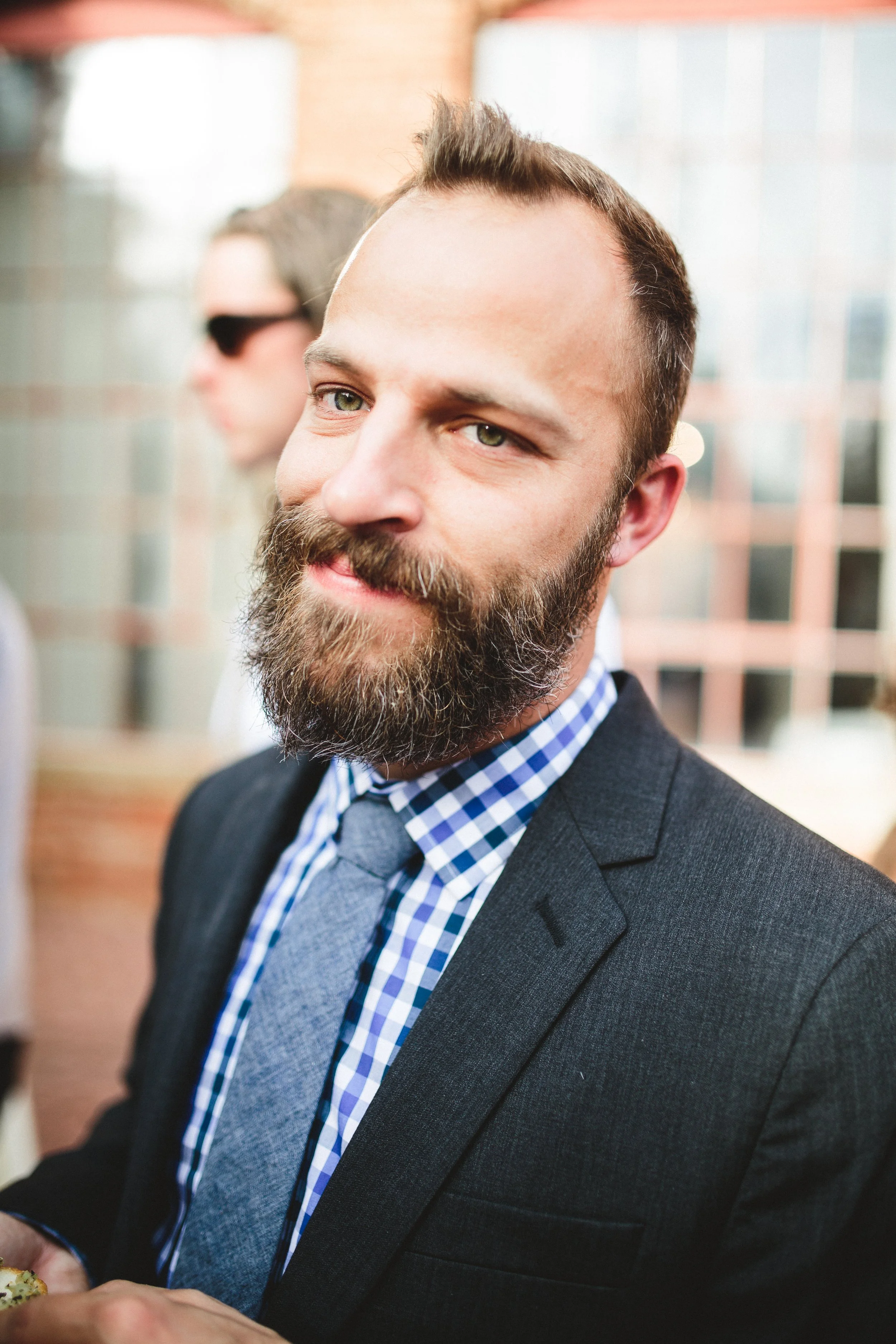 A man with a beard and mustache dressed in a black suit, blue checkered shirt, and gray tie at a formal event, with a woman in sunglasses in the background. It's Chris Jones.