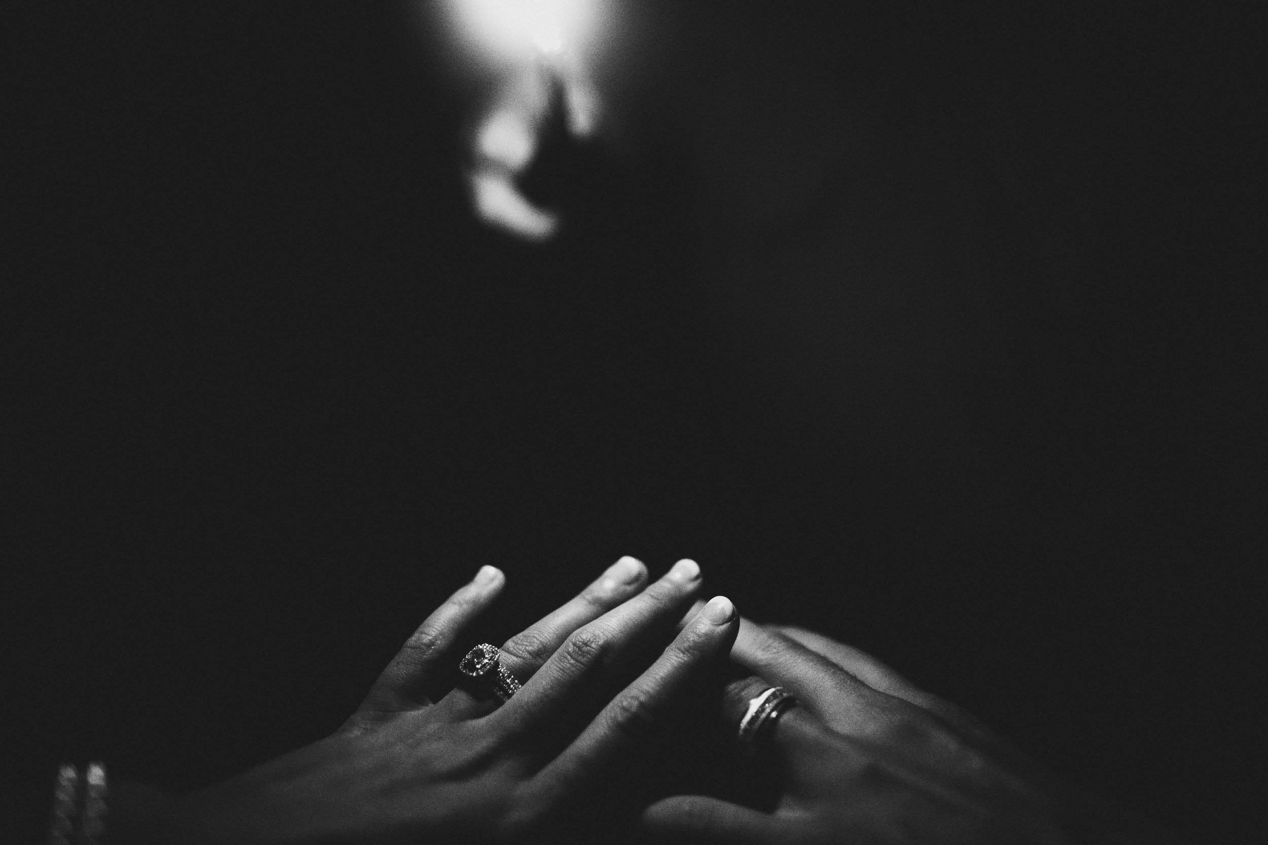 Close-up of two hands with rings, one hand resting on top of the other, against a dark background with a blurred face in the distance. Wedding Photography in Nashville by Sowing Clover Photography