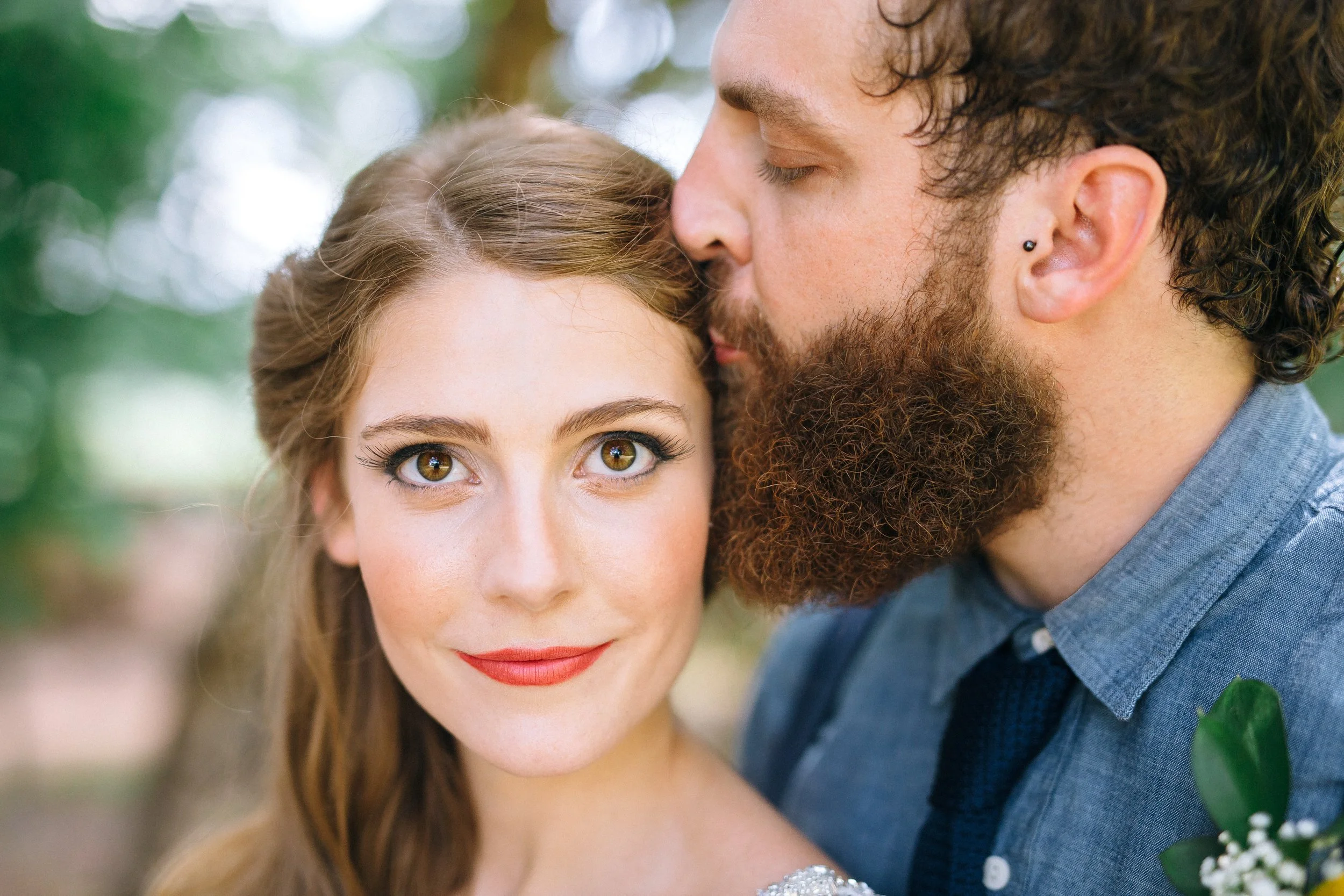 A groom with curly hair and a beard kisses a bride on the forehead. The bride has brown hair, hazel eyes, and red lipstick at Founders Dairy Barn at the Anne Springs Close Greenway South Carolina. Wedding Photography by Sowing Clover Photography.