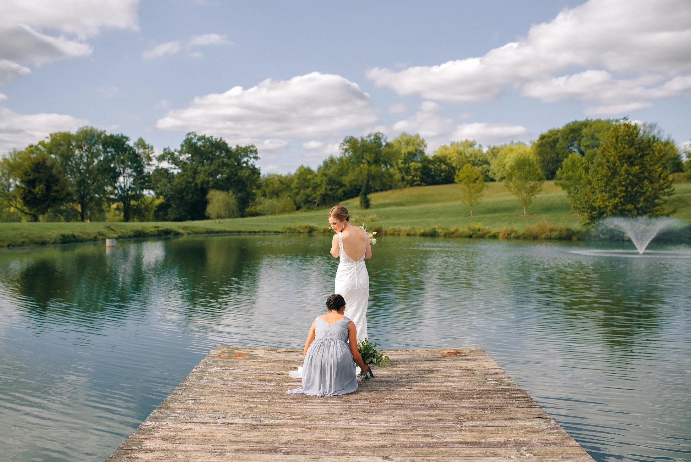 A bride in a white wedding dress standing on a wooden dock by a lake, with a woman in a gray dress kneeling beside her, setting up a floral arrangement at Mint Springs Farm in Tennessee. Wedding photography by Sowing Clover Photography.