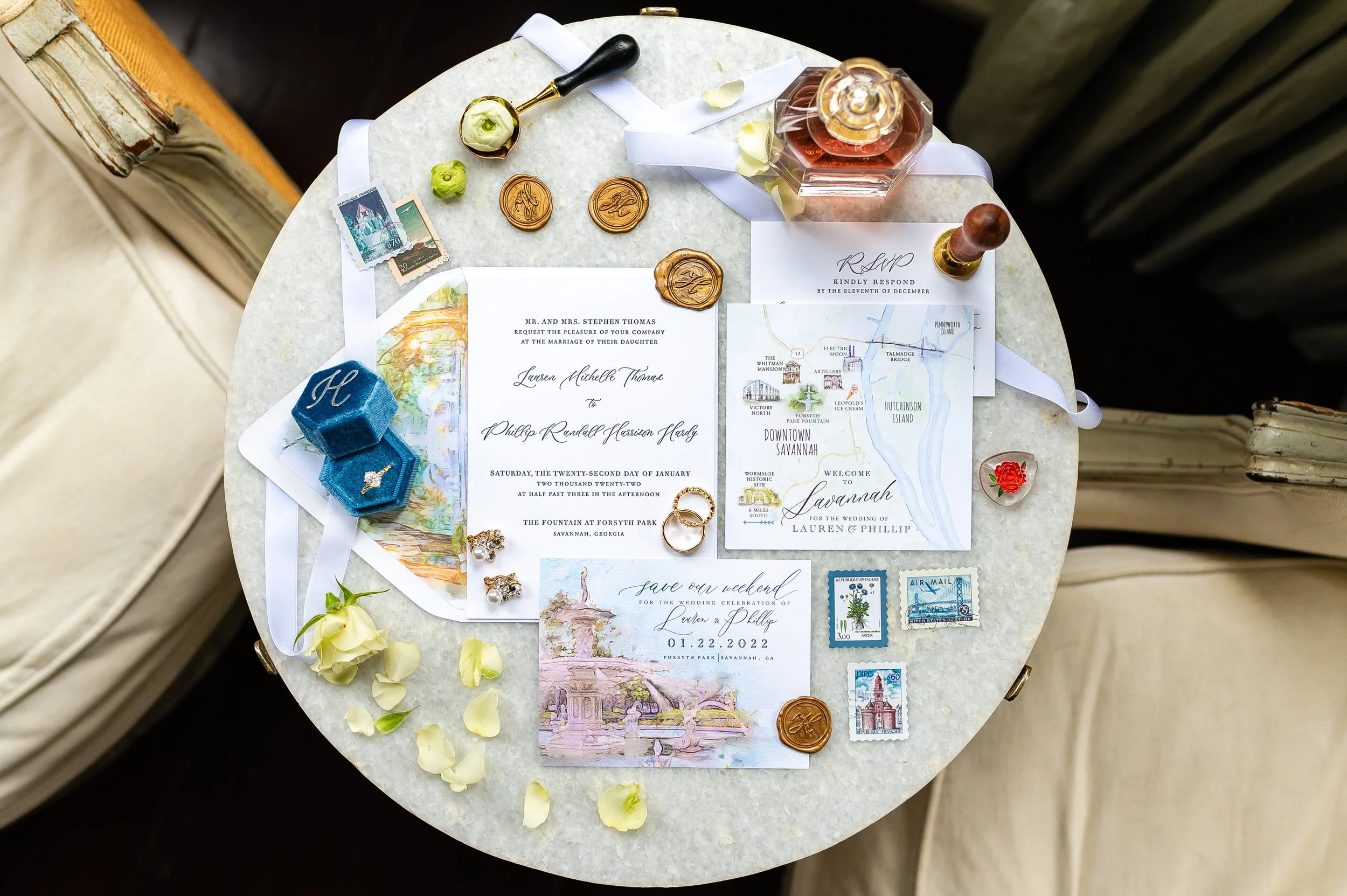Wedding invitation and related items on a white marble table, including wedding invitations, postage stamps, wedding rings, a bottle of perfume, wax seals, a map of Savannah, flowers. Savannah Georgia Wedding photography by Sowing Clover Photography