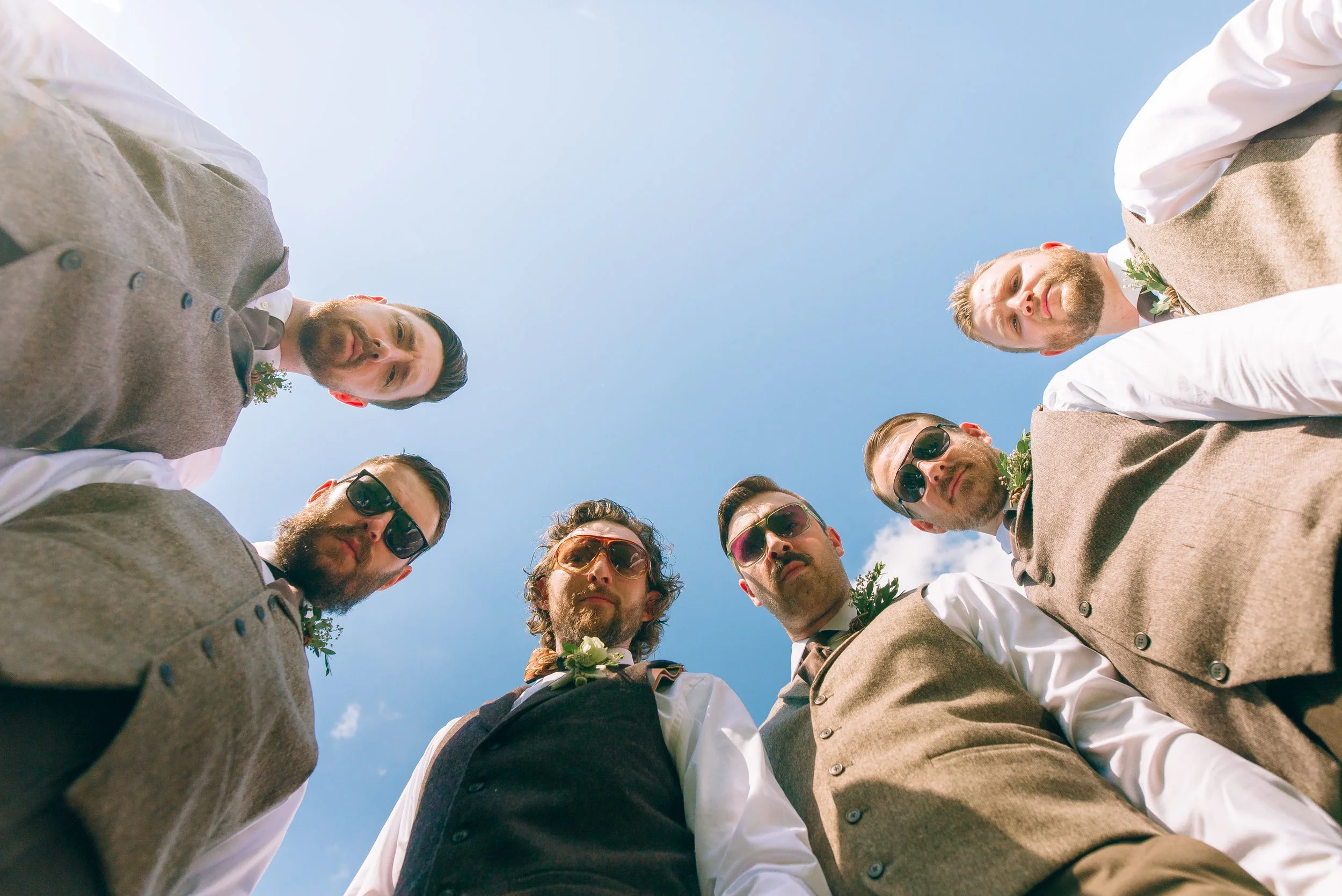 Group of men dressed in formal attire, standing in a circle outdoors, looking down at the camera with a blue sky and some clouds visible above. Atlanta wedding. Wedding Photography by Sowing Clover Photography.