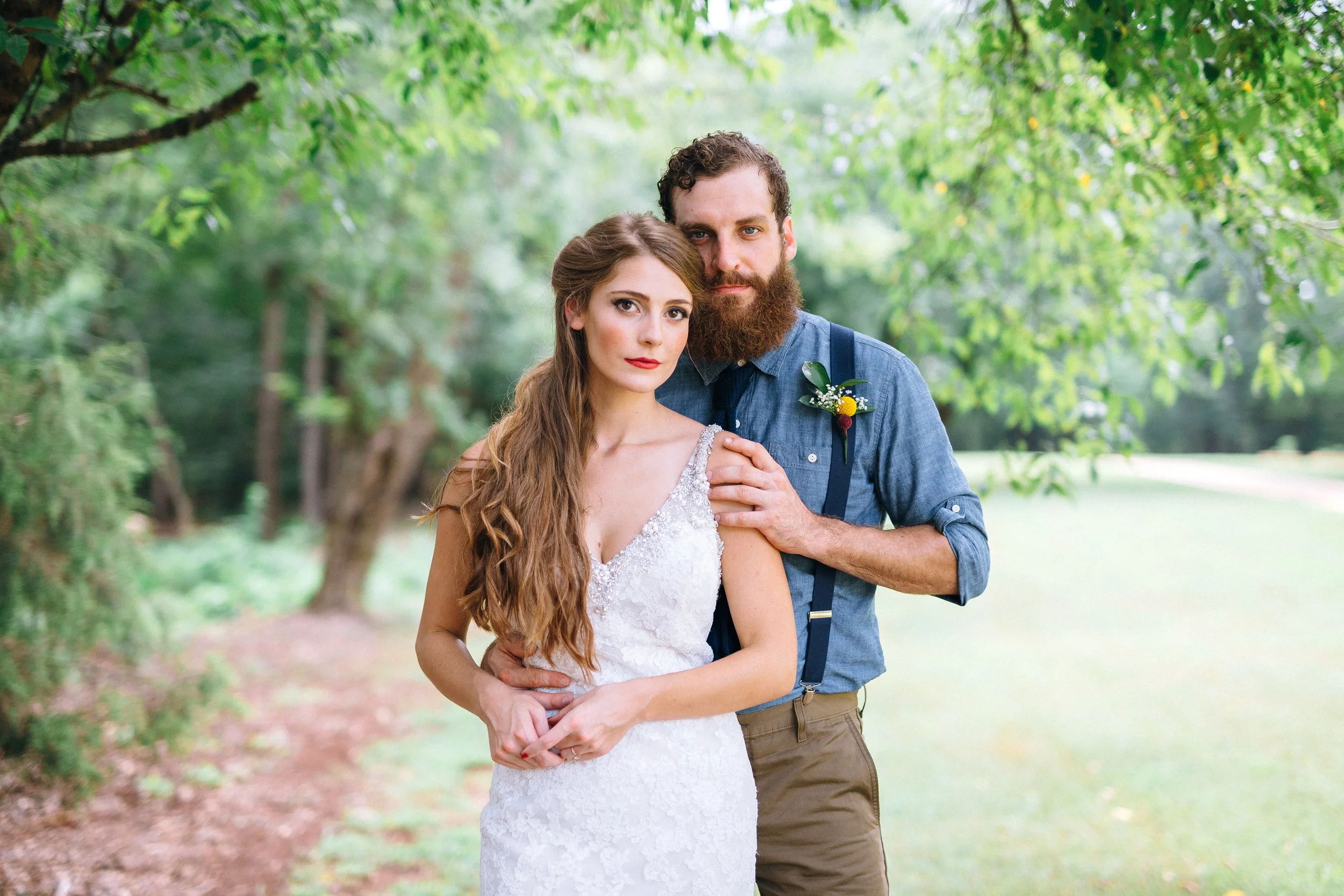 A bride and groom couple standing close together outdoors amidst green trees, with the woman wearing a white lace wedding dress at Founders Dairy Barn at the Anne Springs Close Greenway South Carolina. Wedding Photography by Sowing Clover Photography