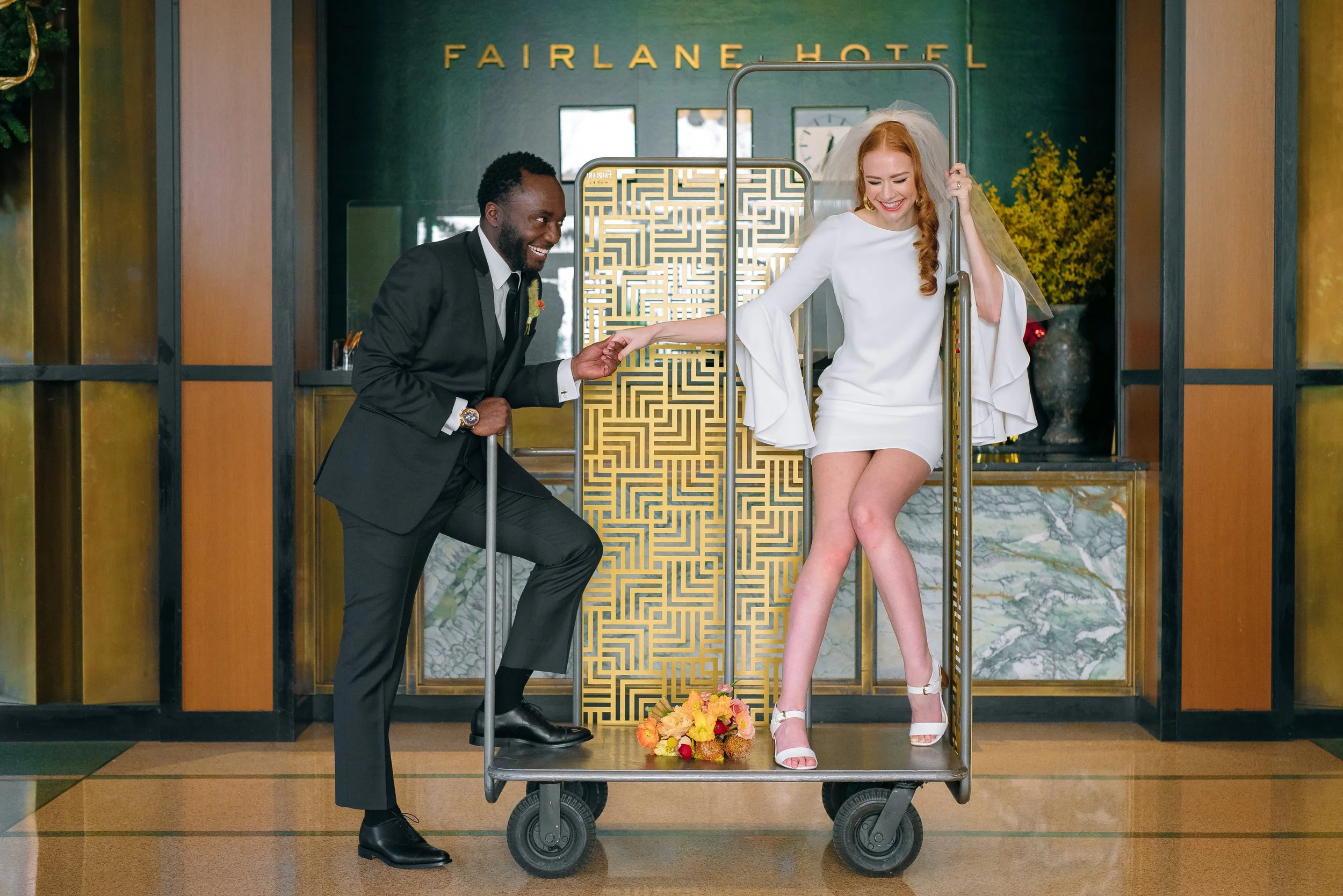 A bride in a white wedding dress and high heels is riding a luggage cart with flowers on the floor at the Fairlane Hotel. A man in a suit is pushing the cart, holding her hand, and smiling. In Nashville Wedding photography by Sowing Clover Photograph