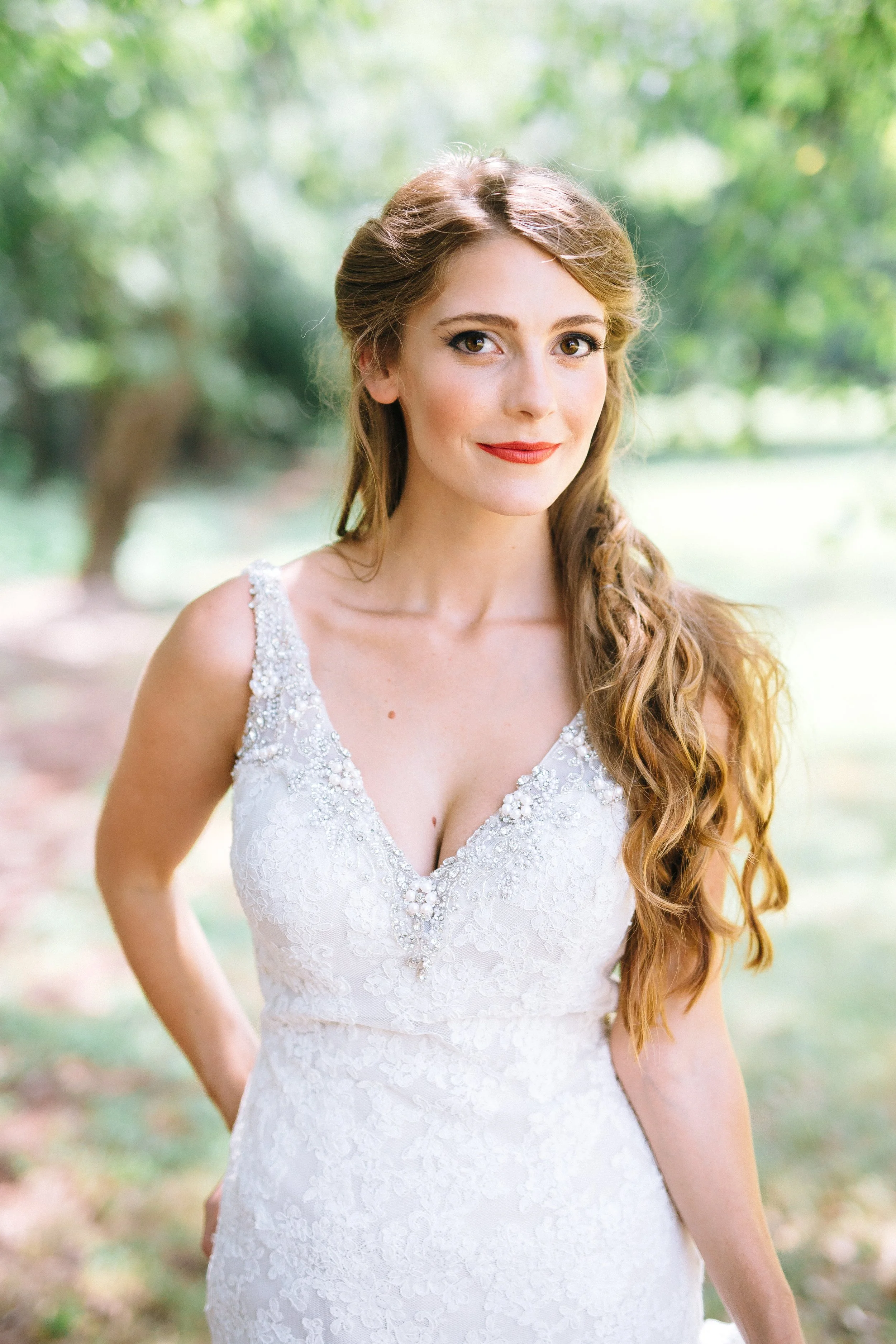 A bride with long, wavy red hair in a white wedding dress with bead and pearl embellishments, standing outdoors with trees at Founders Dairy Barn at the Anne Springs Close Greenway South Carolina. Wedding Photography by Sowing Clover Photography.