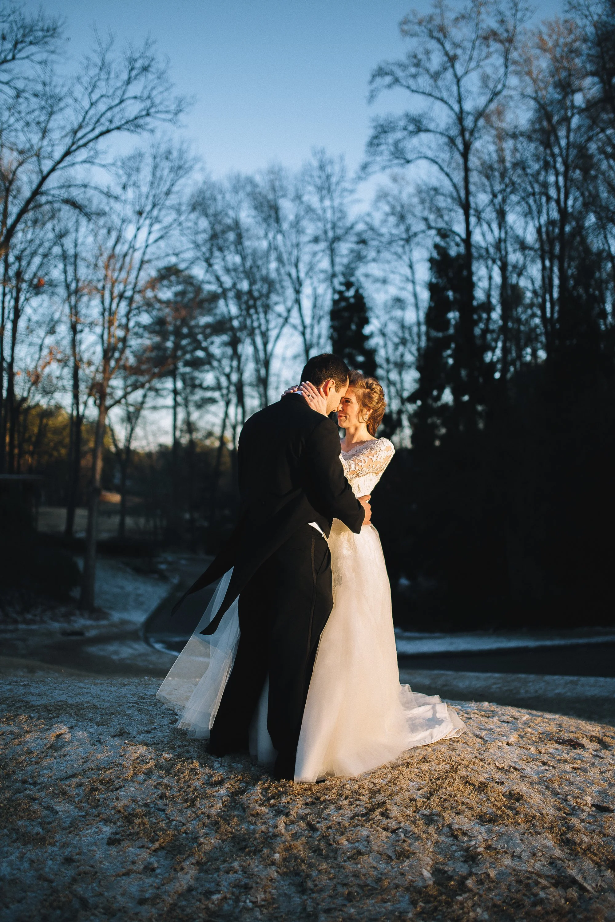 A bride and groom embrace outdoors during twilight, with leafless trees and a cold, snowy ground in the background. Country Club of the South Wedding Photography. photography by Sowing Clover Photography.