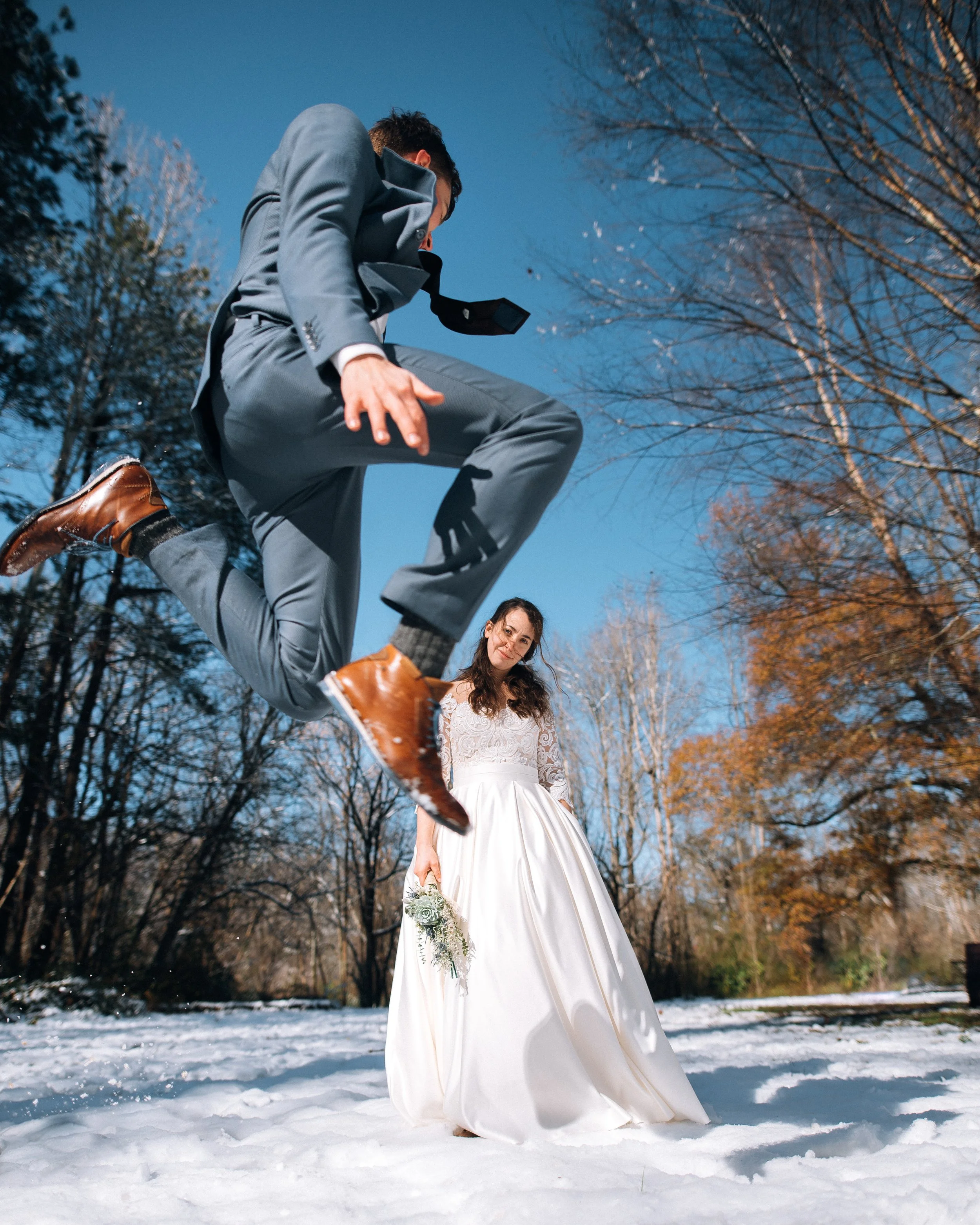A groom in a gray suit jumping in the air in a snowy outdoor setting with a bride in a wedding dress standing in the snow and holding a bouquet.  Snowy Atlanta wedding. Wedding photography by Sowing Clover Photography