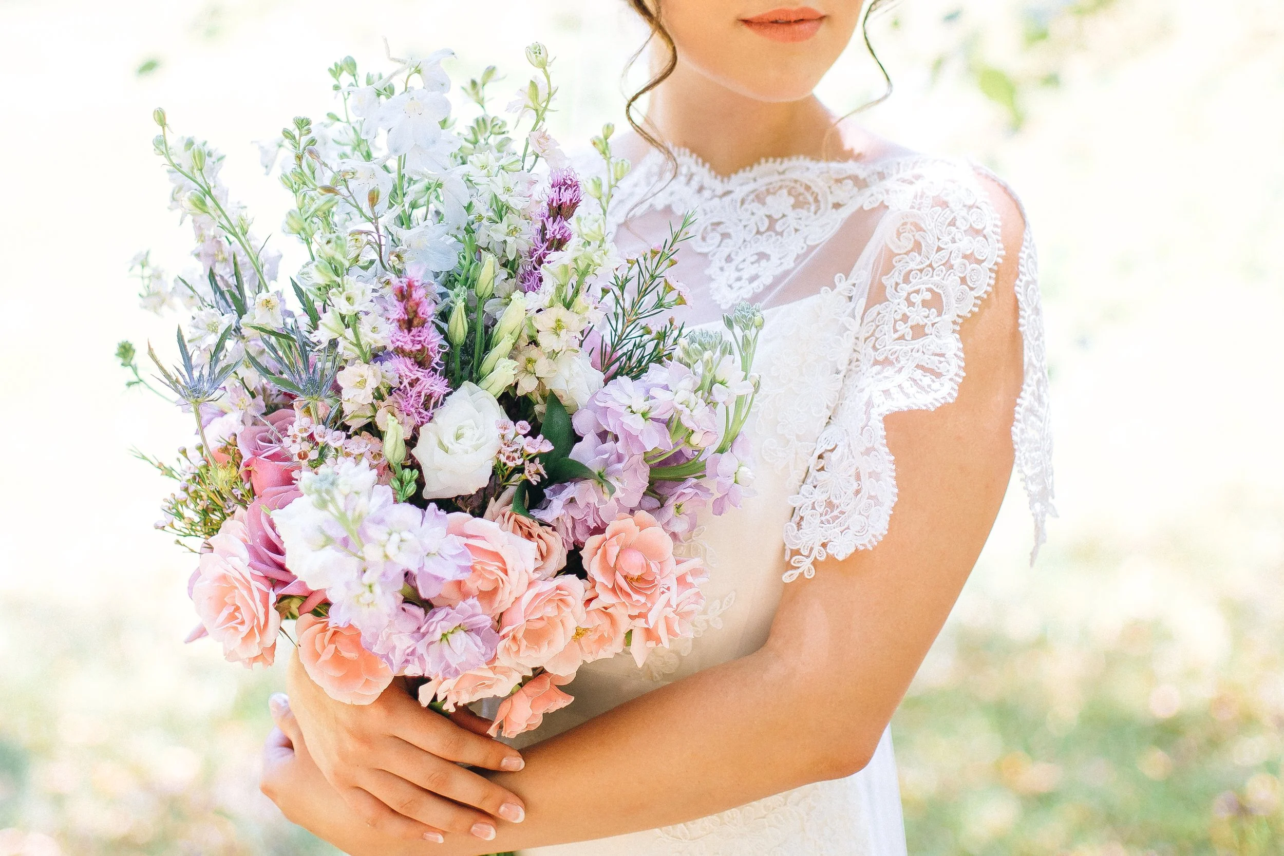 A bride holding a bouquet of pink, purple, white, and pastel-colored flowers, wearing a white lace dress, outdoors with sunlight in Atlanta for a wedding. Wedding Photography in by Sowing Clover Photography