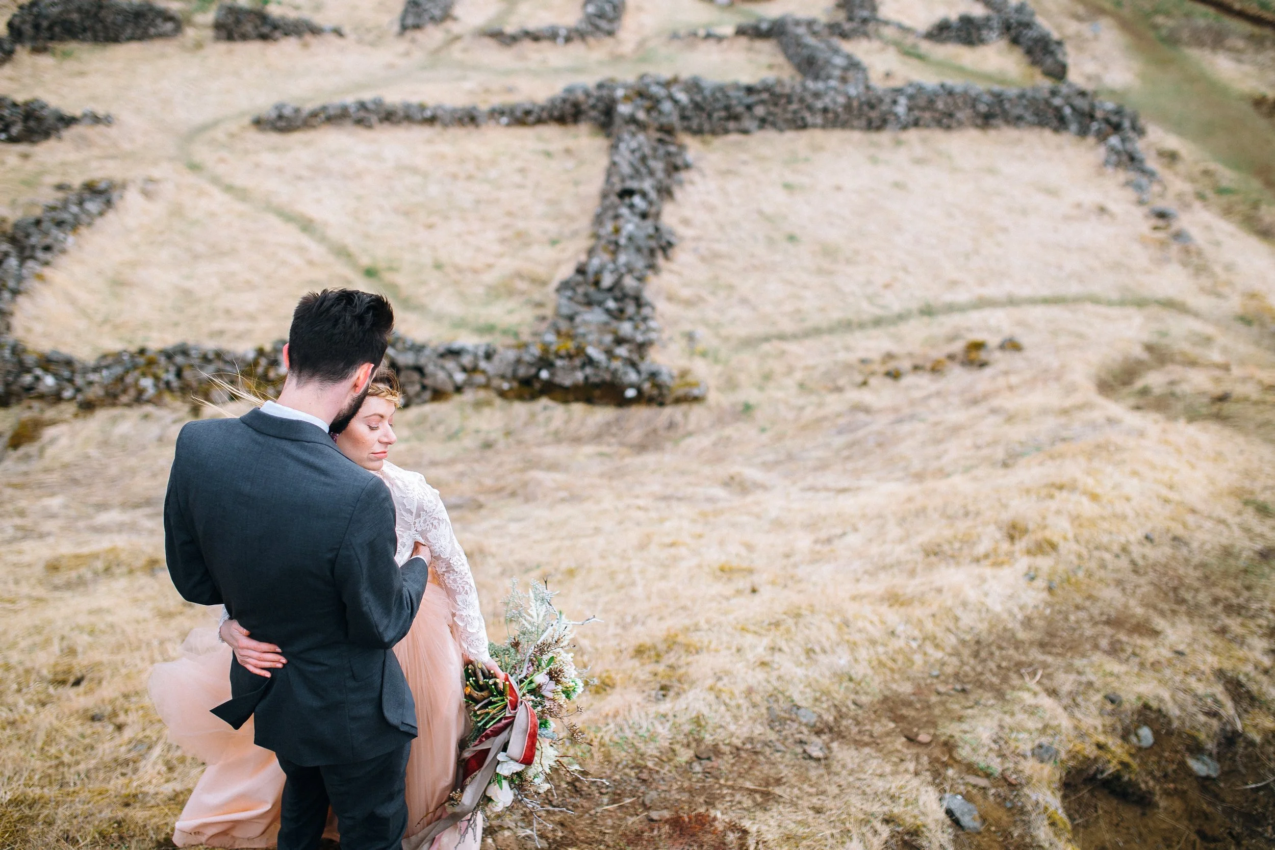 A man and a woman dressed in a suit and a dress are embracing outdoors, with a dry stone wall and grassy hillside in the background  in Iceland. Wedding Photography by Sowing Clover Photography..