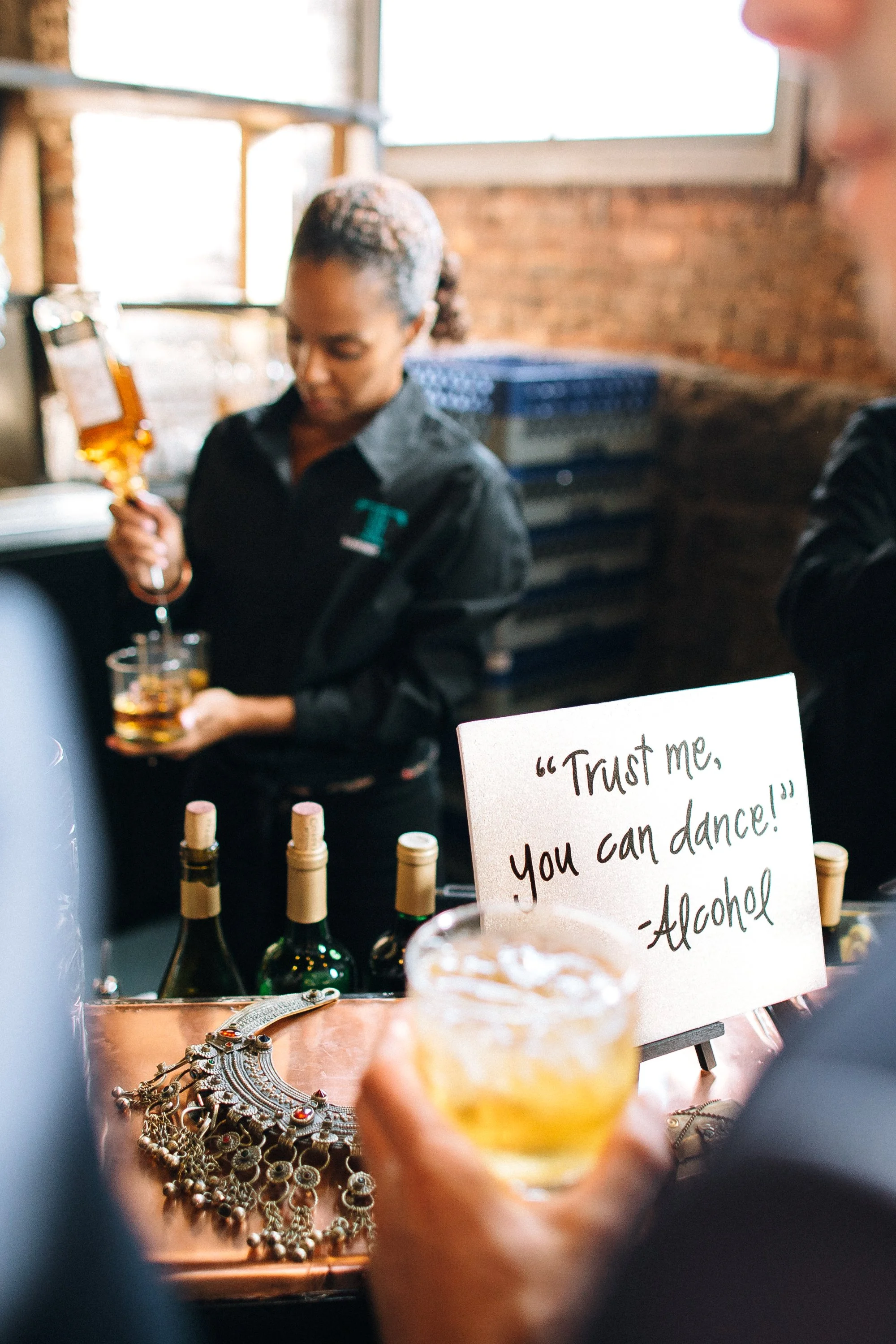 A bartender pouring alcohol into a glass. A person in the foreground is holding a drink and there is a sign that reads "Trust me, you can dance!" - Alcohol. Atlanta Wedding photography by Sowing Clover Photography