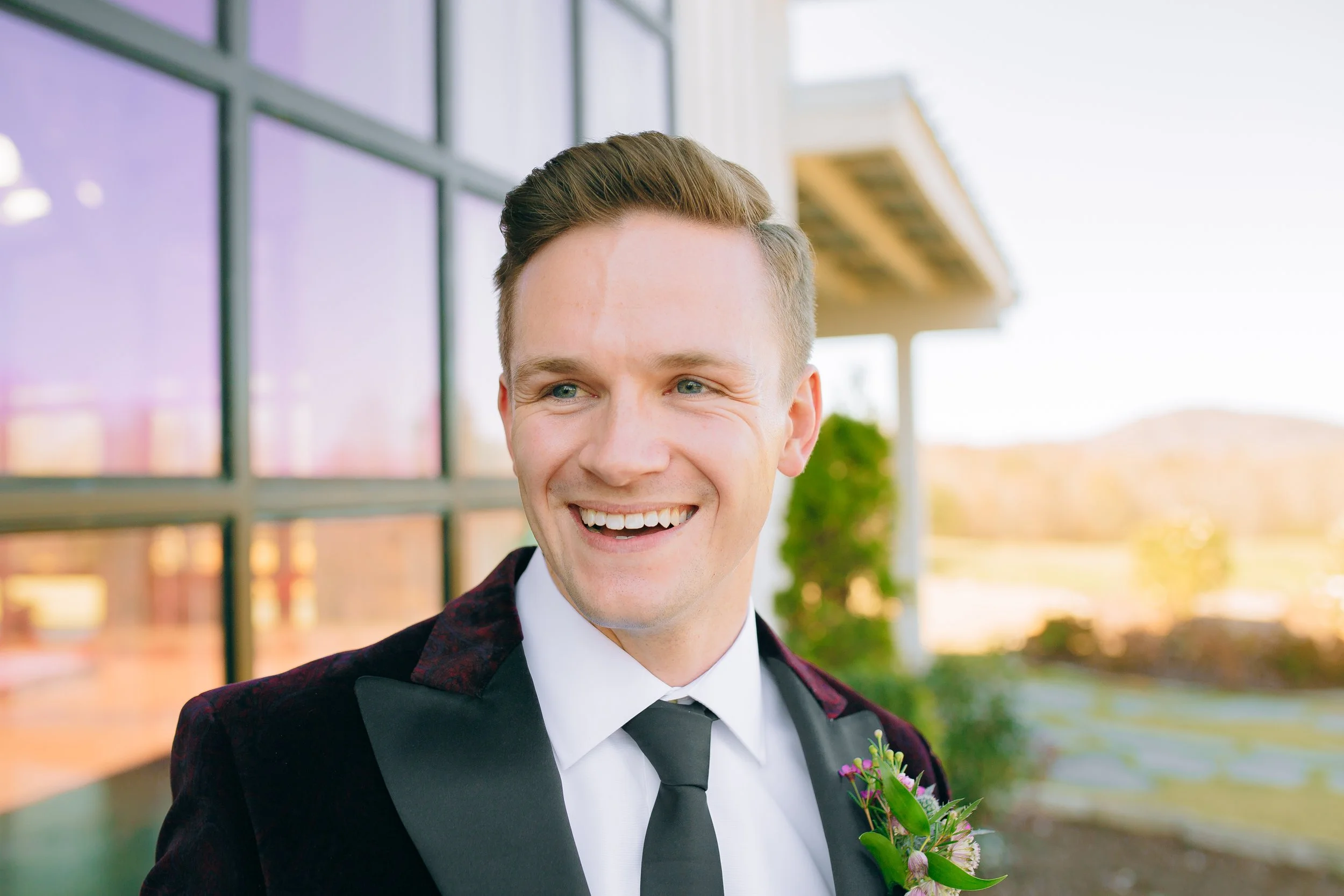 Young groom in formal attire smiling outdoors with glass building and mountains in the background at a north Georgia wedding.  Wedding Photography by Sowing Clover Photography.
