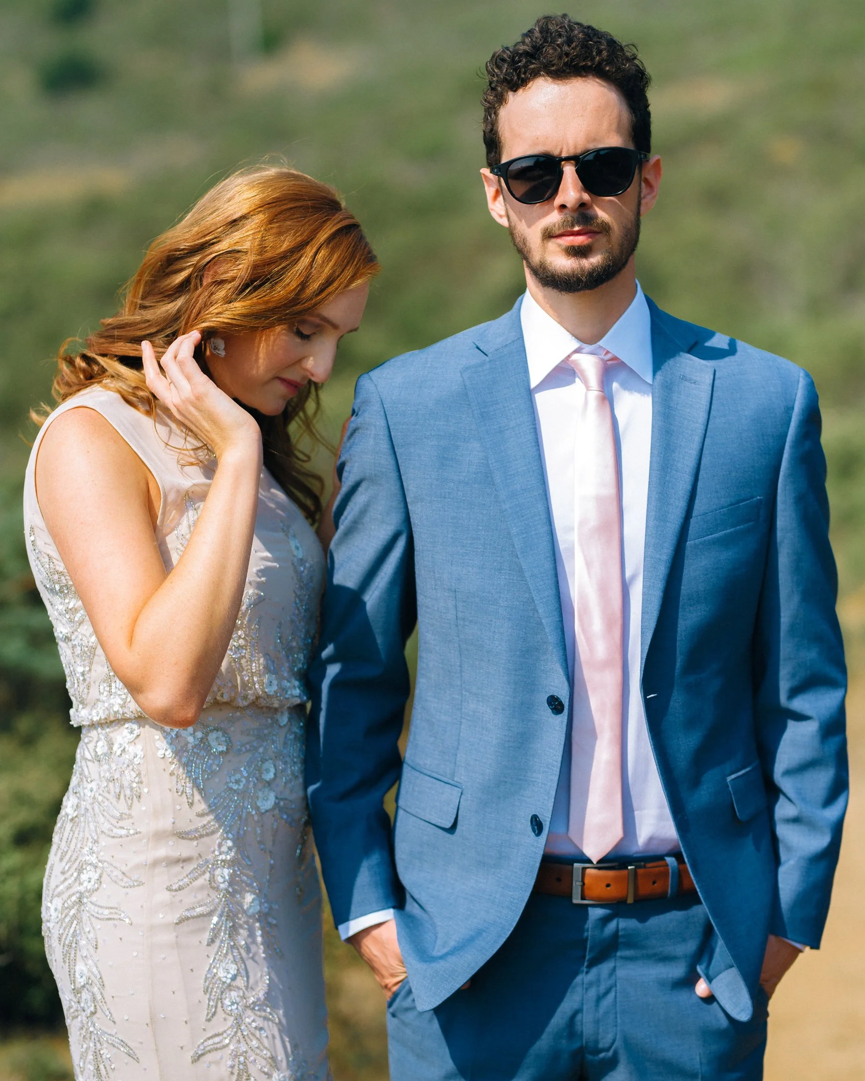 A groom wearing a blue suit, sunglasses, and a pink tie stands outdoors with a bride in a light-colored, embroidered wedding dress, who is touching her hair and looking down in San Fransisco. Wedding Photography by Sowing Clover Photography.