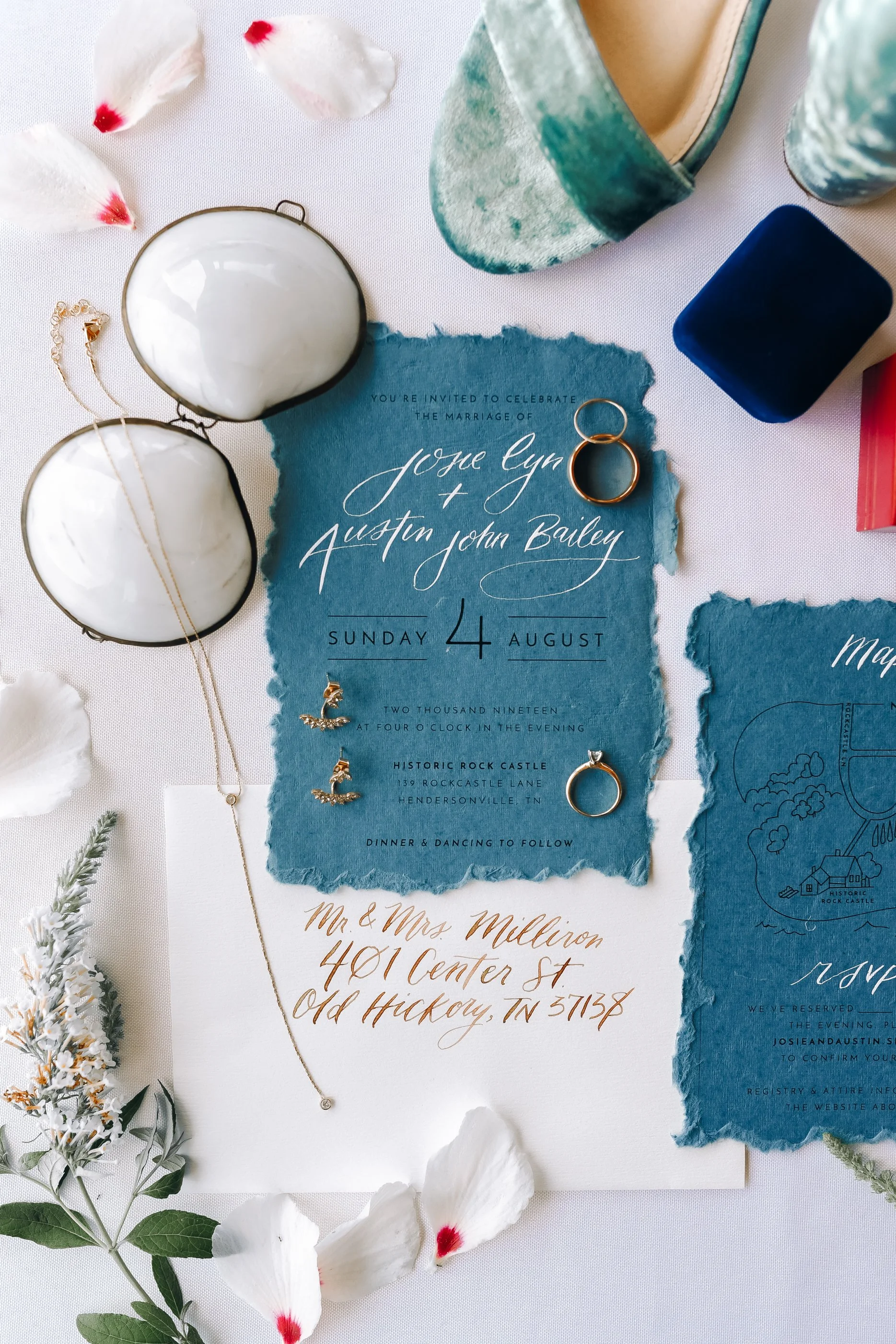 Wedding invitation with rings, jewelry, and decorative flowers on a white surface, featuring torn blue paper with wedding details  at Historic Rock Castle in Hendersonville. Wedding Photography by Sowing Clover Photography.