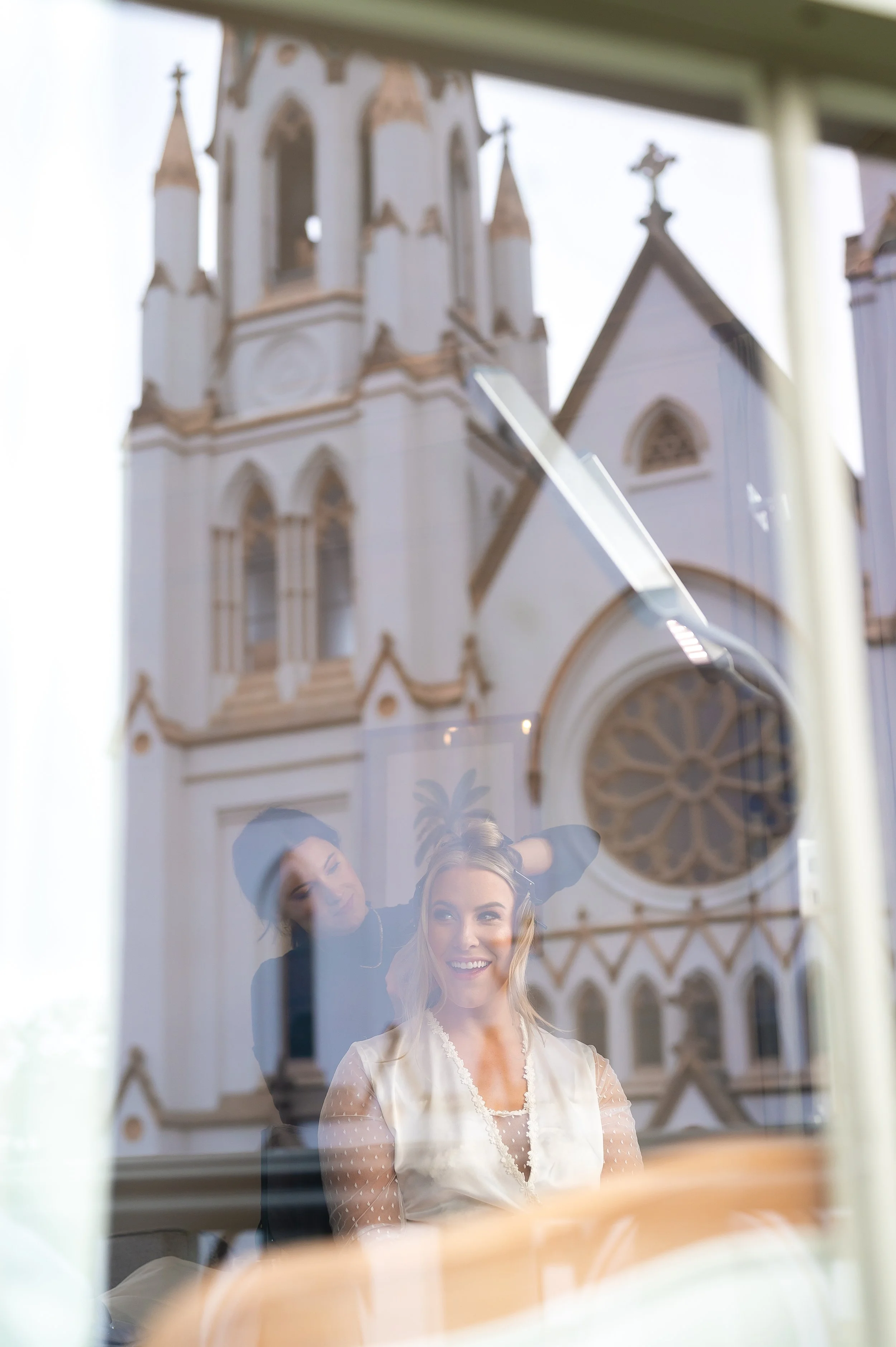 A woman smiling as she gets her hair styled by a hairstylist, reflected in a window with the backdrop The Cathedral Basilica of St. John the Baptist in Savannah Georgia.  Wedding Photography by Sowing Clover Photography.