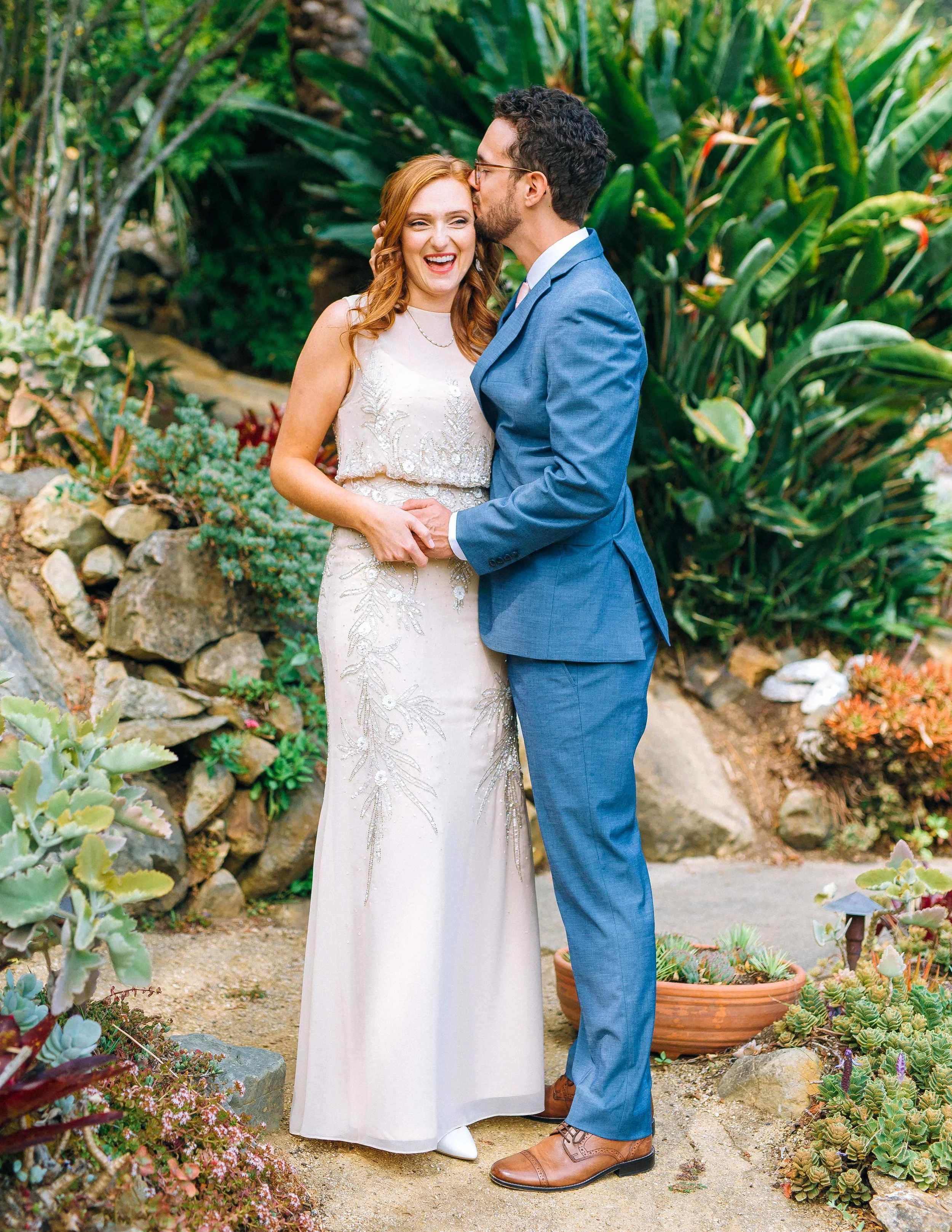 A newlywed couple in wedding attire holding hands and sharing a kiss in a lush garden setting in San Francisco. Wedding photography by Sowing Clover Photography.