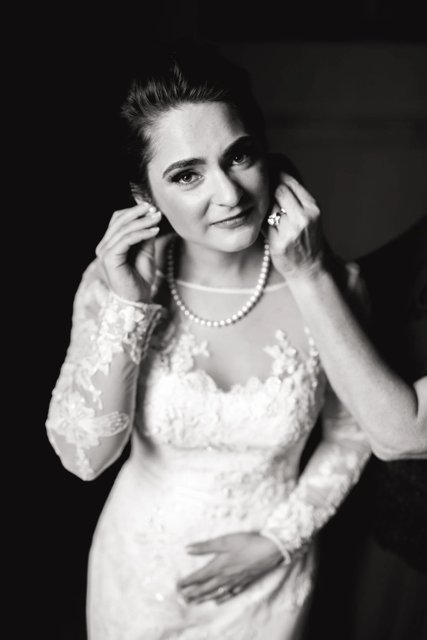 A black and white photo of a woman in a lace wedding dress, wearing a pearl necklace, adjusting her earrings, smiling slightly, with one hand on her waist in North Georgia. Wedding Photography by Sowing Clover Photography.